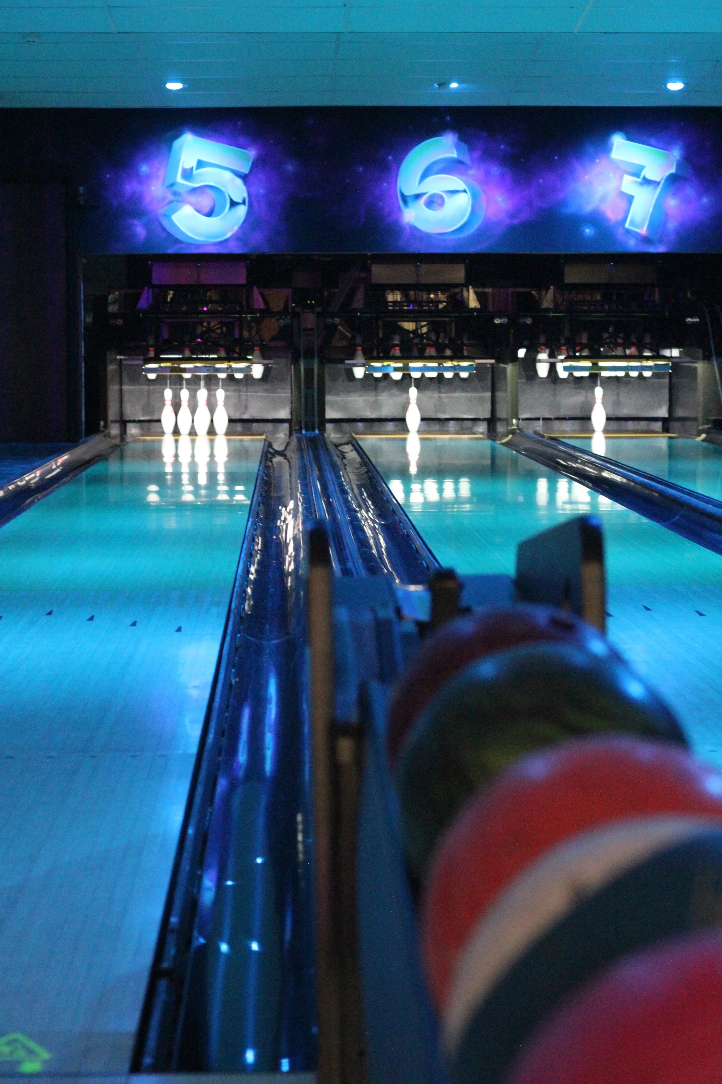 A bowling alley with bowling pins set up at the end of the lane, illuminated by blue lighting. Large illuminated numbers '5', '6', and '7' are displayed on a sign above the lane.