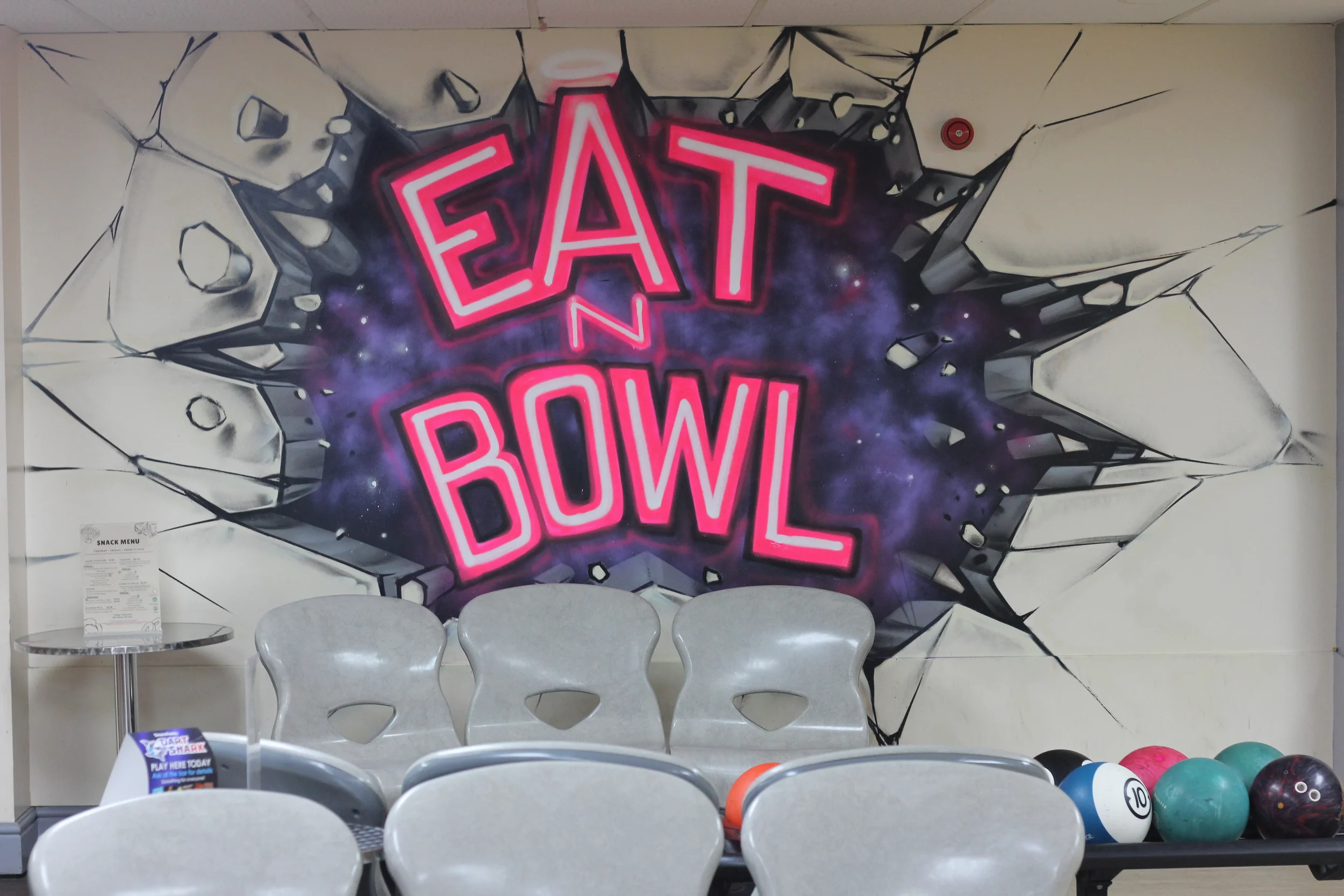 Colorful mural with the text "Eat and Bowl" in neon pink letters, depicting a galaxy background with graffiti-style cracks, located in a bowling alley with chairs and bowling balls.