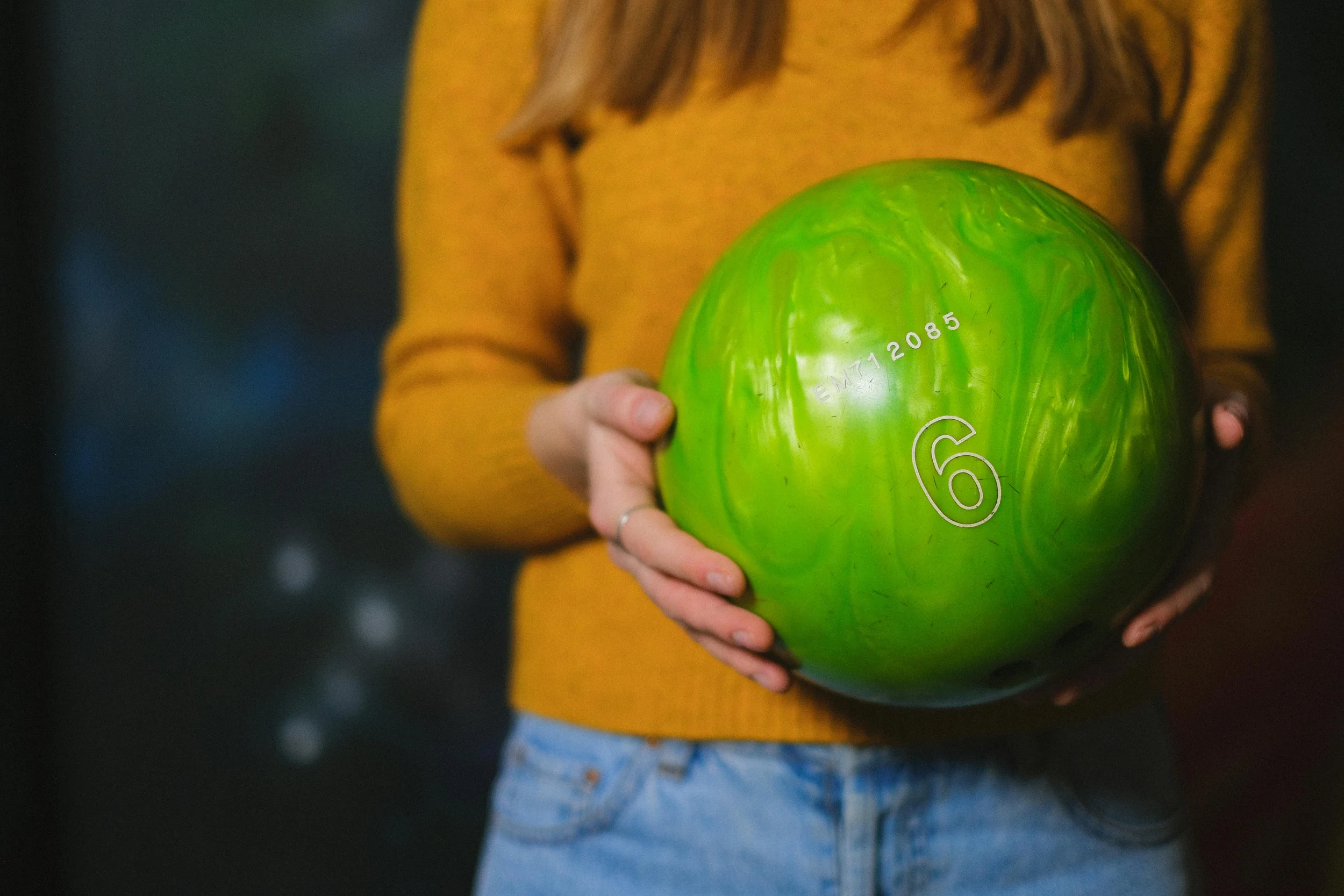 Person wearing a yellow sweater and blue jeans holding a green bowling ball.