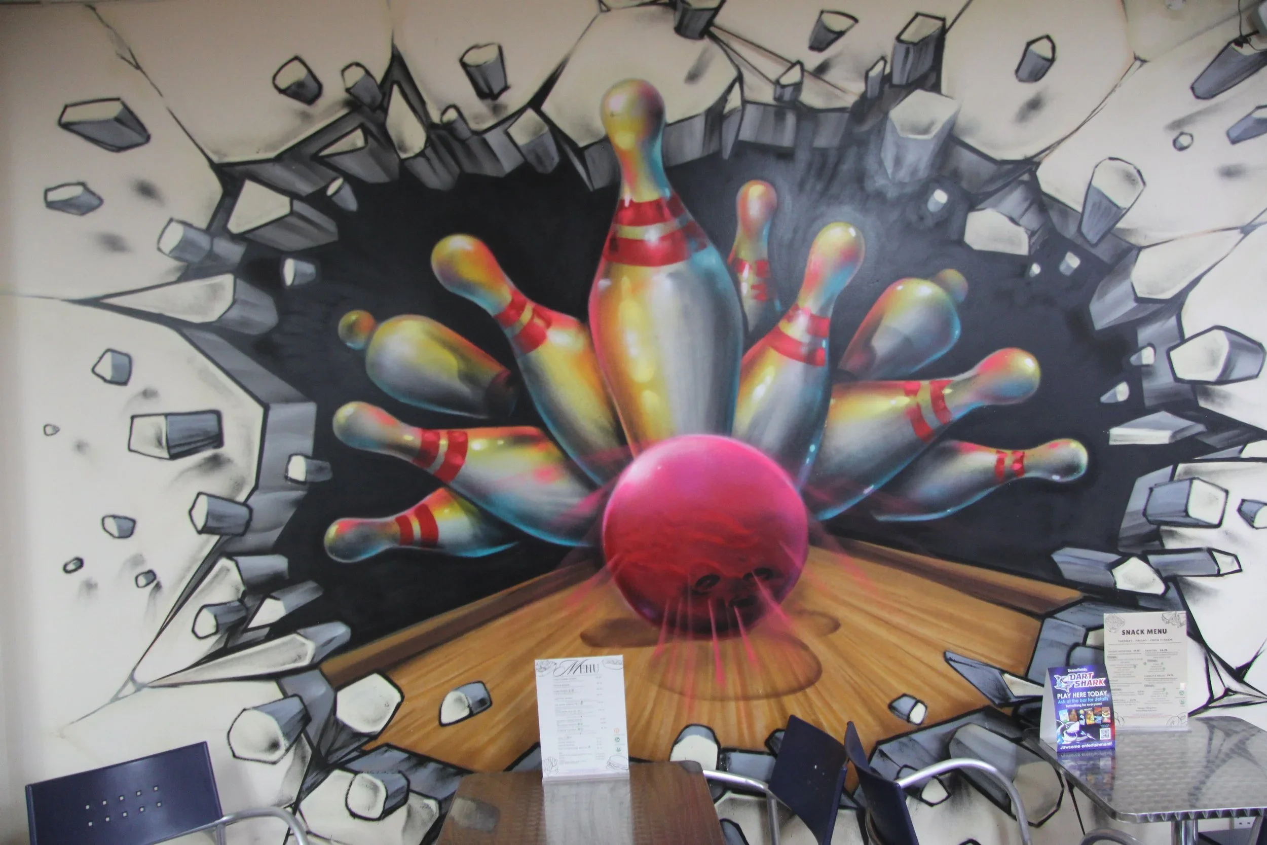 A mural of colorful bowling pins crashing through a black wall, creating a visual effect of the pins breaking through the wall and hitting a bowling ball on a wooden lane.