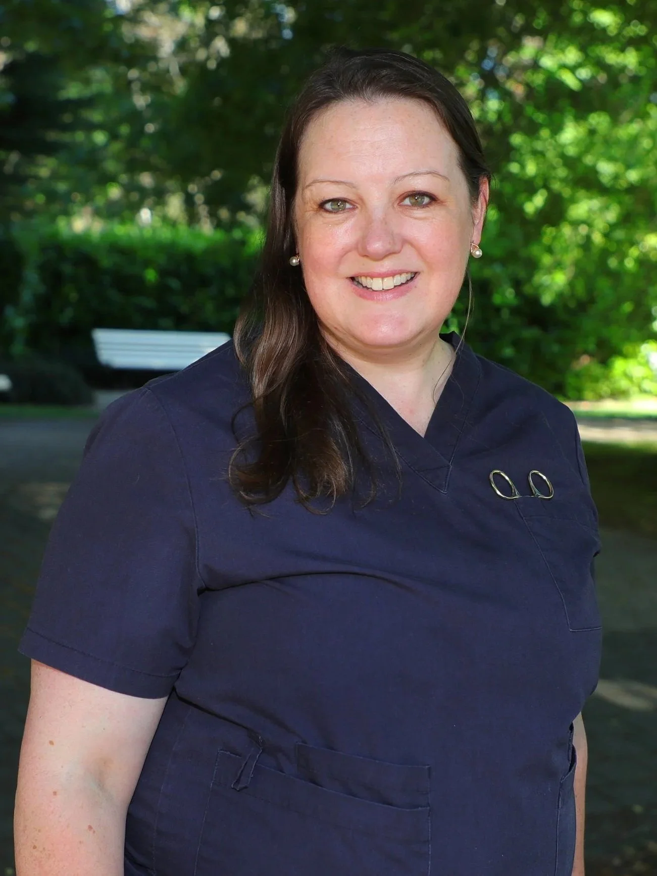 A woman with long brown hair wearing a dark navy medical uniform, standing outdoors with green foliage in the background.