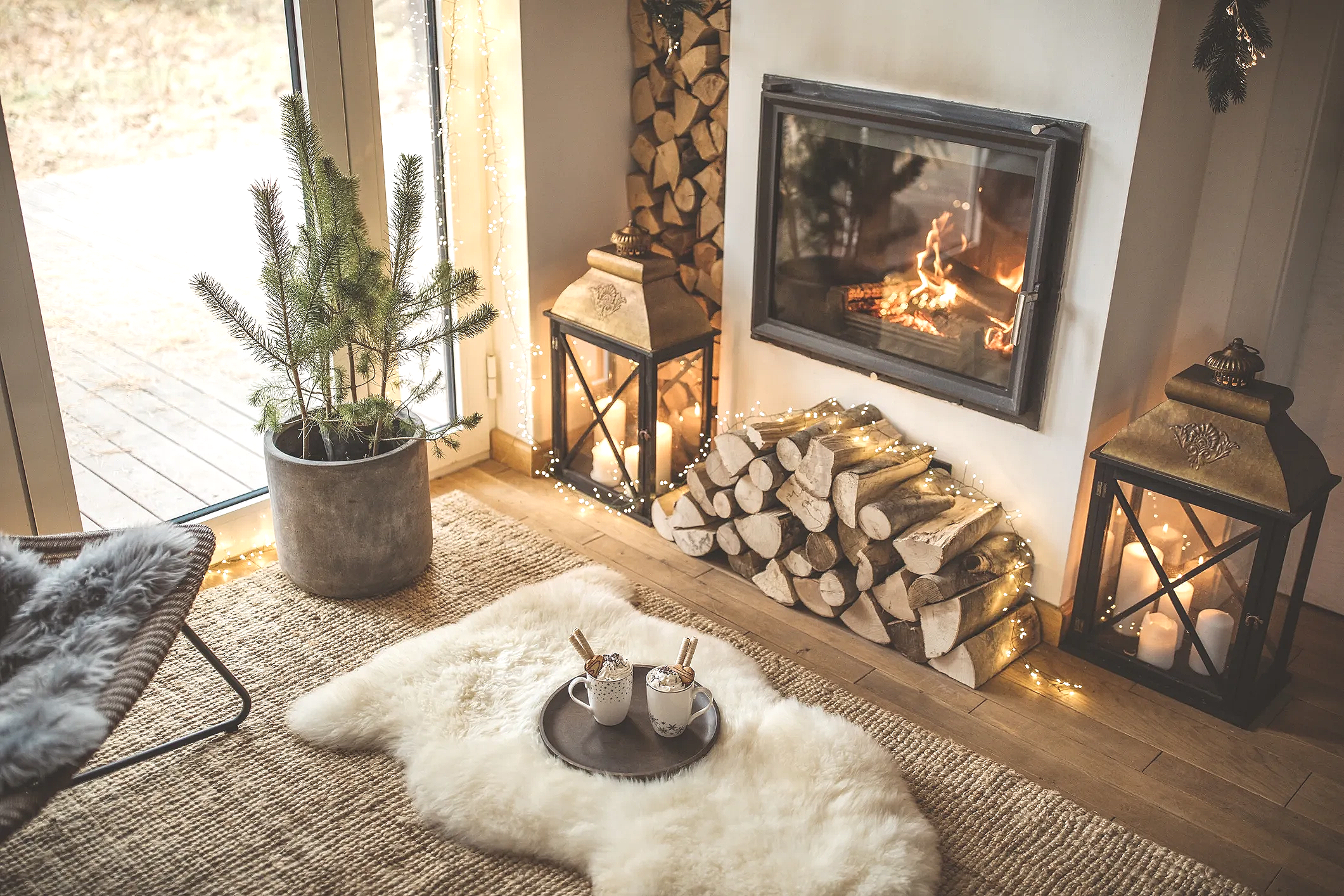 Cozy living room with fire in a fireplace, stacked firewood, two lanterns with candles, a potted pine tree, a sheepskin rug with two mugs of hot chocolate and marshmallows, and a sliding glass door leading outside.