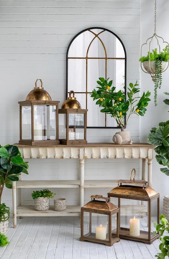 Decorative interior with a white distressed console table, green plants, gold lanterns, candles, and a window mirror, all set against a white brick wall.