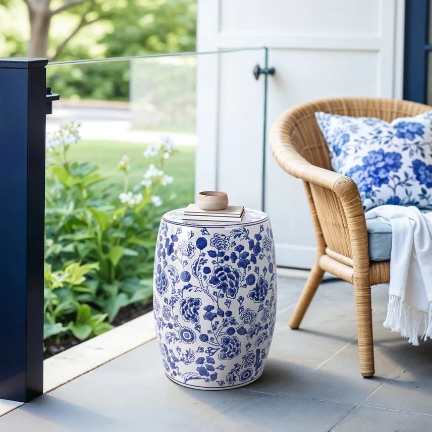  porch with a woven chair, a blue and white pillow, a small ceramic cup on a striped notebook, and a ceramic garden stool with blue floral patterns, overlooking a garden with green plants.