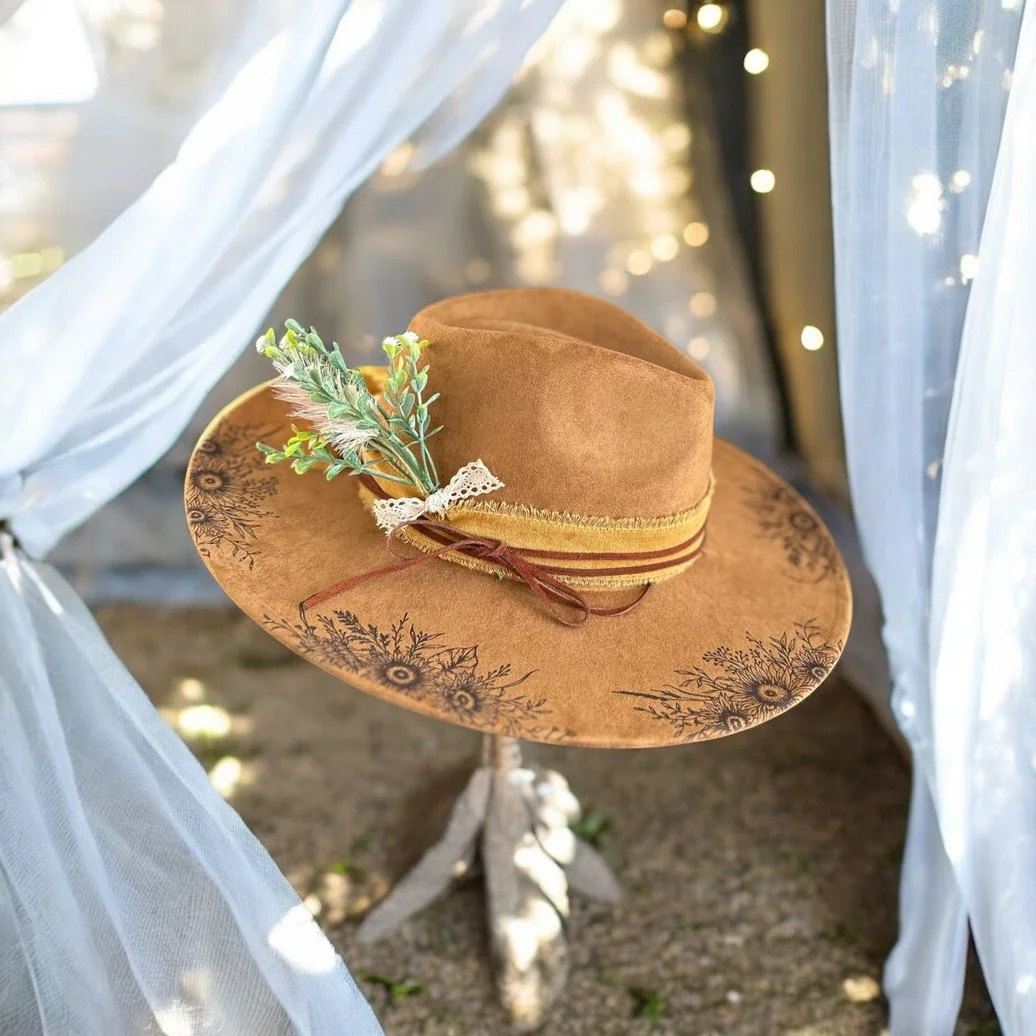 A brown cowboy hat with decorative floral patterns around the brim, placed on a wooden stand or chair, with white sheer curtains framing the scene and blurred string lights in the background.
