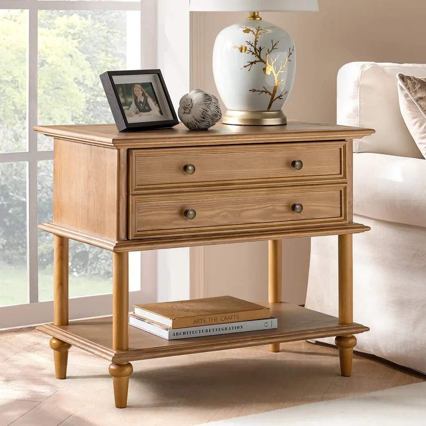 Light wood nightstand with two drawers, top decorated with a framed photo, a decorative ball, and a white ceramic lamp with gold tree branch design. Bottom shelf holds books. Part of a beige sofa and a sliding glass door are visible.