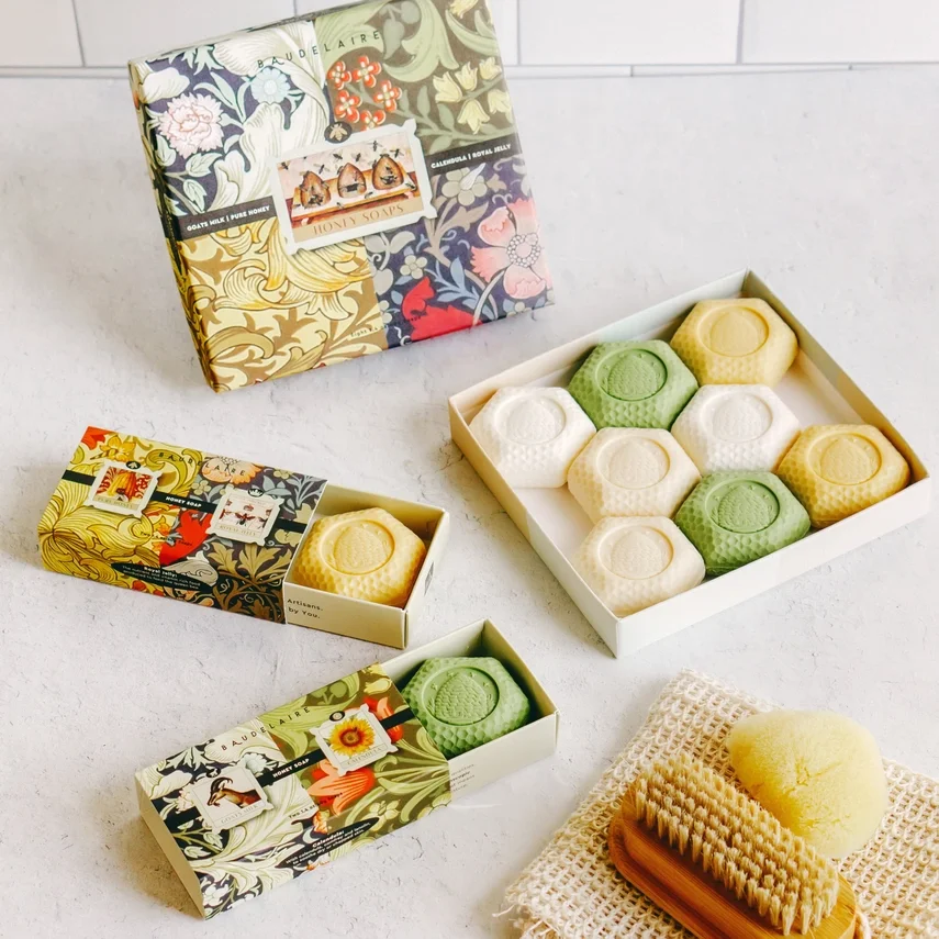 Assorted beeswax skincare products including soap bars, a bath sponge, and a body brush, displayed on a white surface with decorative packaging.