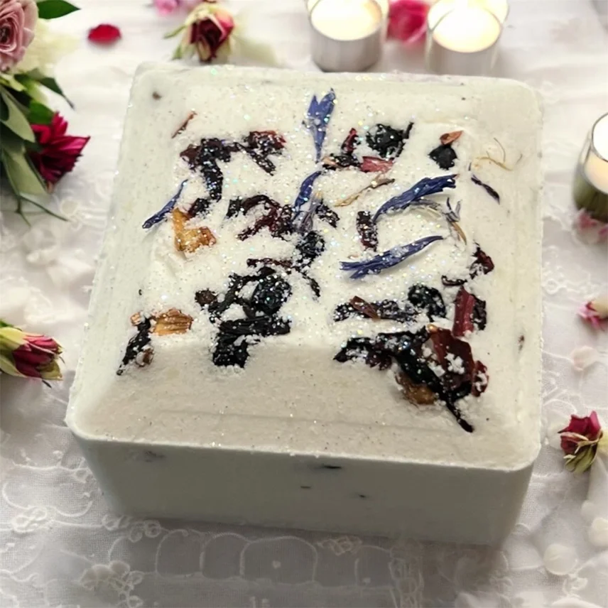 Square white cake with colorful flower petal decorations on top, surrounded by flowers and candles.