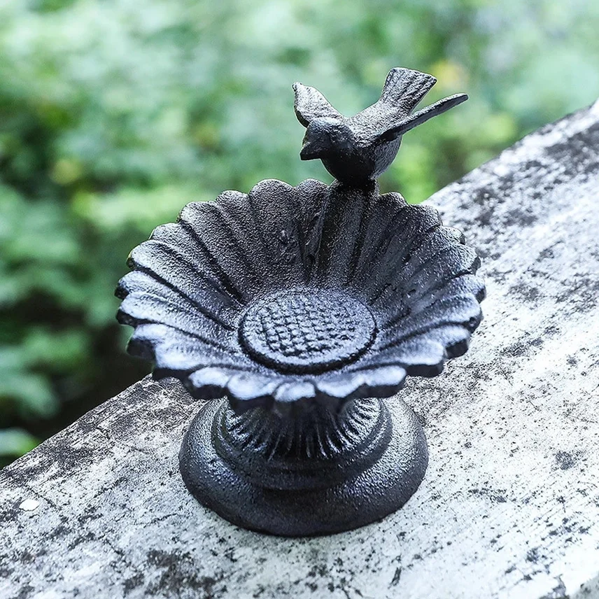Small black cast iron birdbath with a bird figurine on the edge, placed on a weathered surface with blurred green foliage in the background.