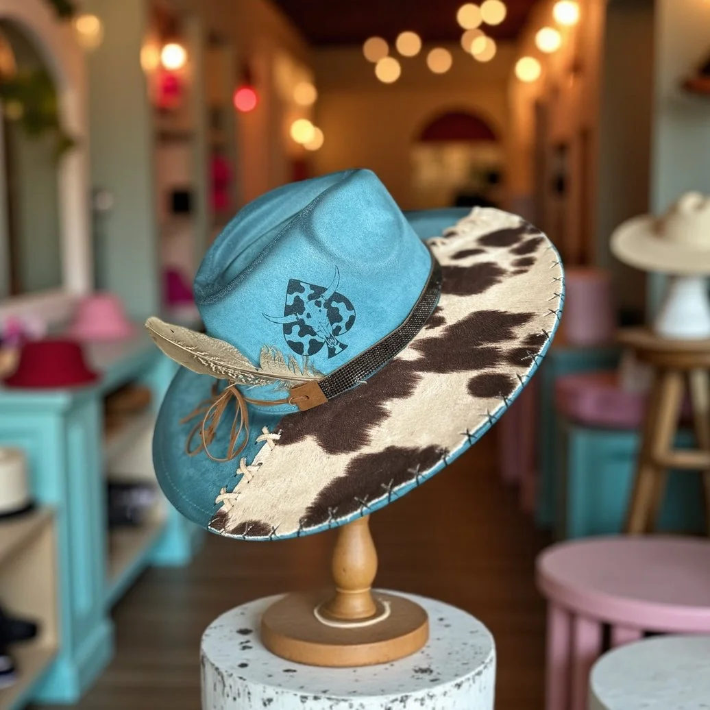 A turquoise cowboy hat with a cow print brim displayed on a wooden stand in a boutique or decorative shop.