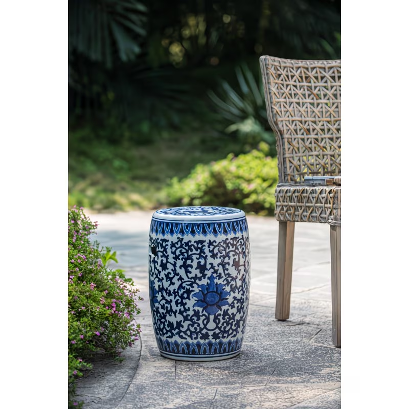 A decorative ceramic stool with intricate blue and white patterns, placed outdoors on a stone patio next to a woven chair and purple flowering bushes, with greenery in the background.