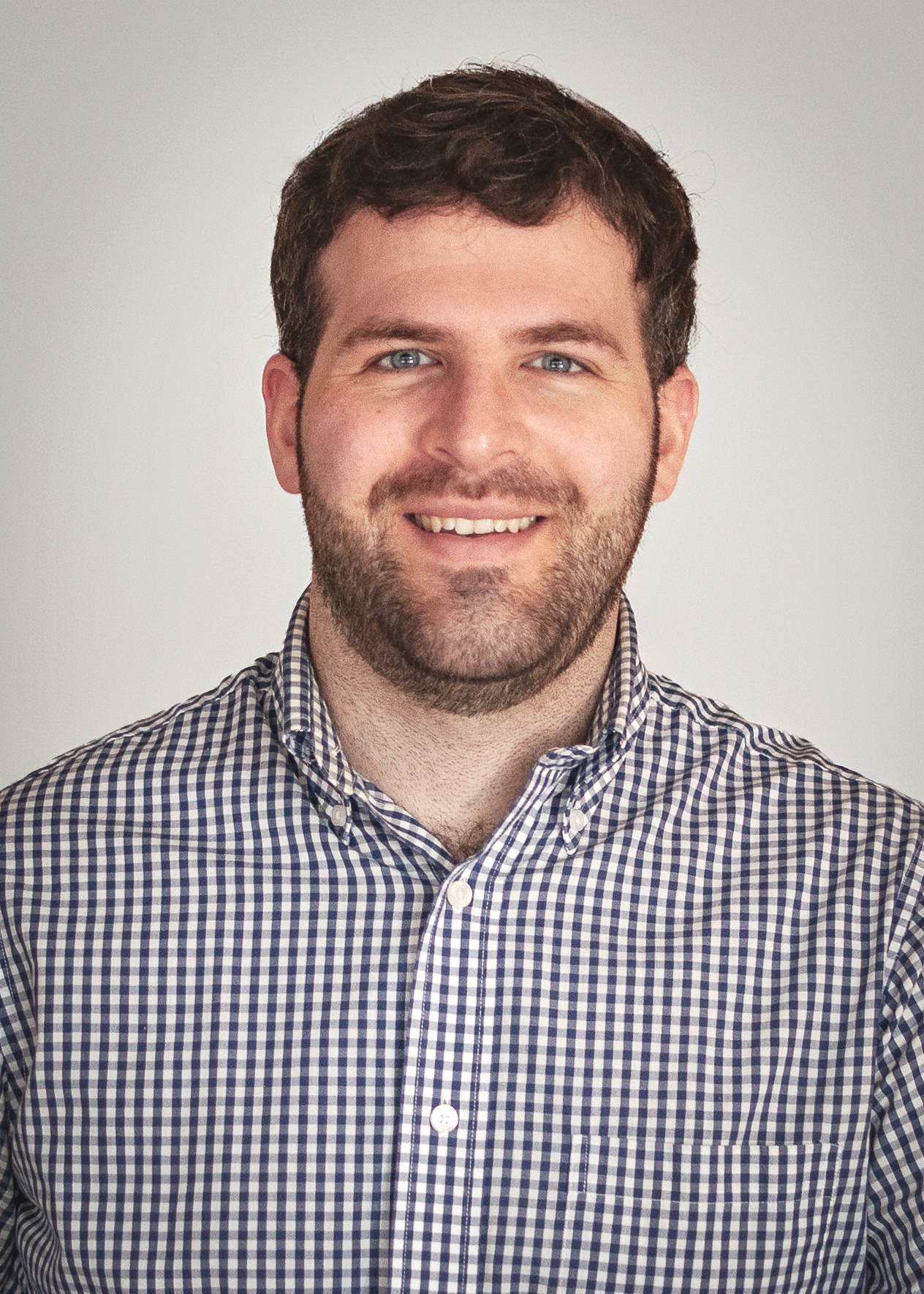 A man with short brown hair, blue eyes, and a beard, smiling, wearing a blue and white checkered button-up shirt, against a plain light-colored background.