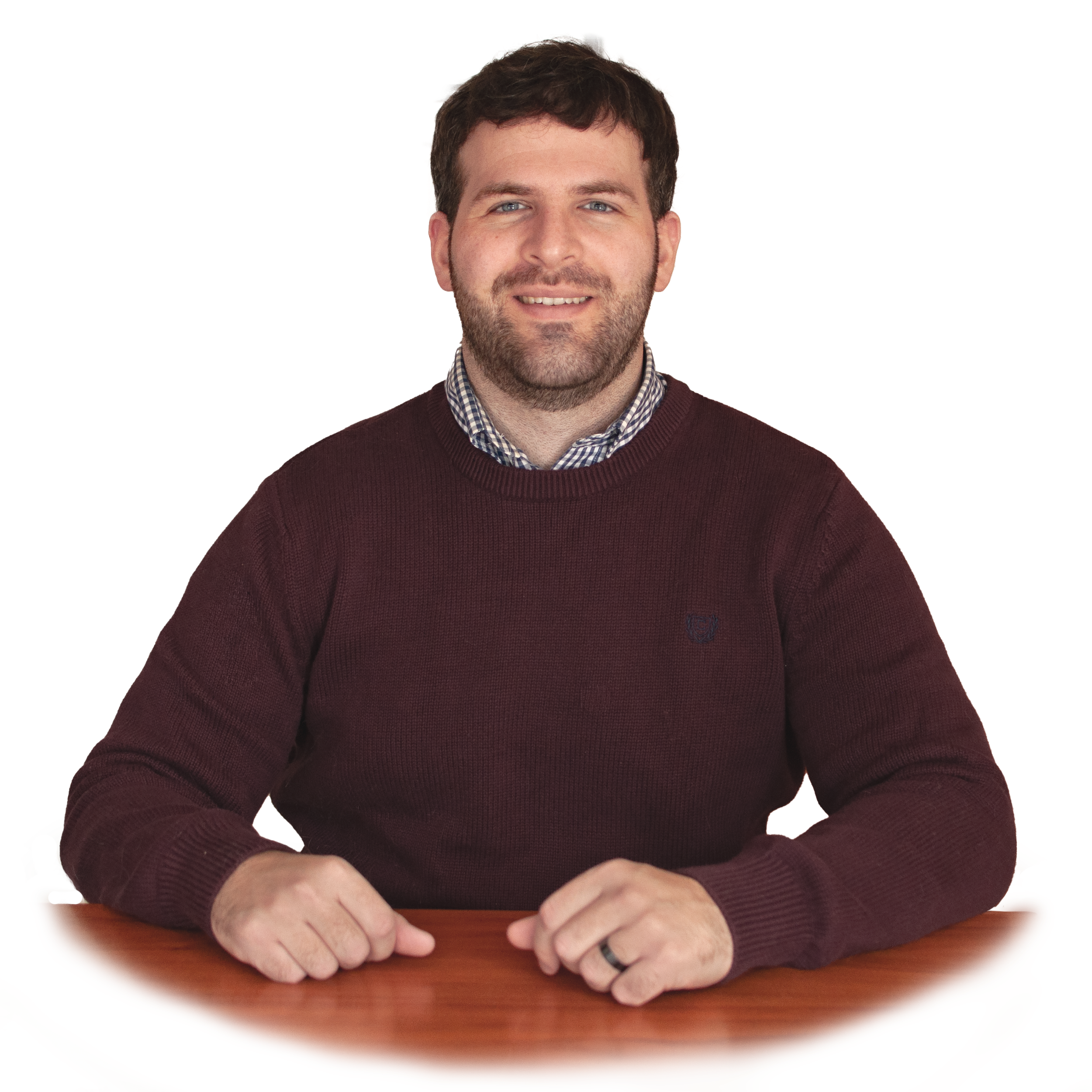 A man with dark hair and a beard sitting at a wooden table, wearing a maroon sweater over a checkered shirt, smiling at the camera.