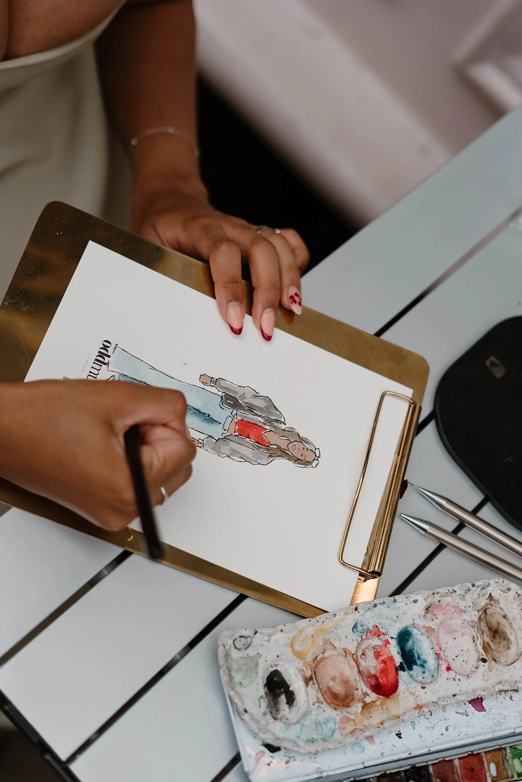 Person drawing a fashion sketch on a clipboard with a marker, surrounded by painting supplies and pens on a white desk.