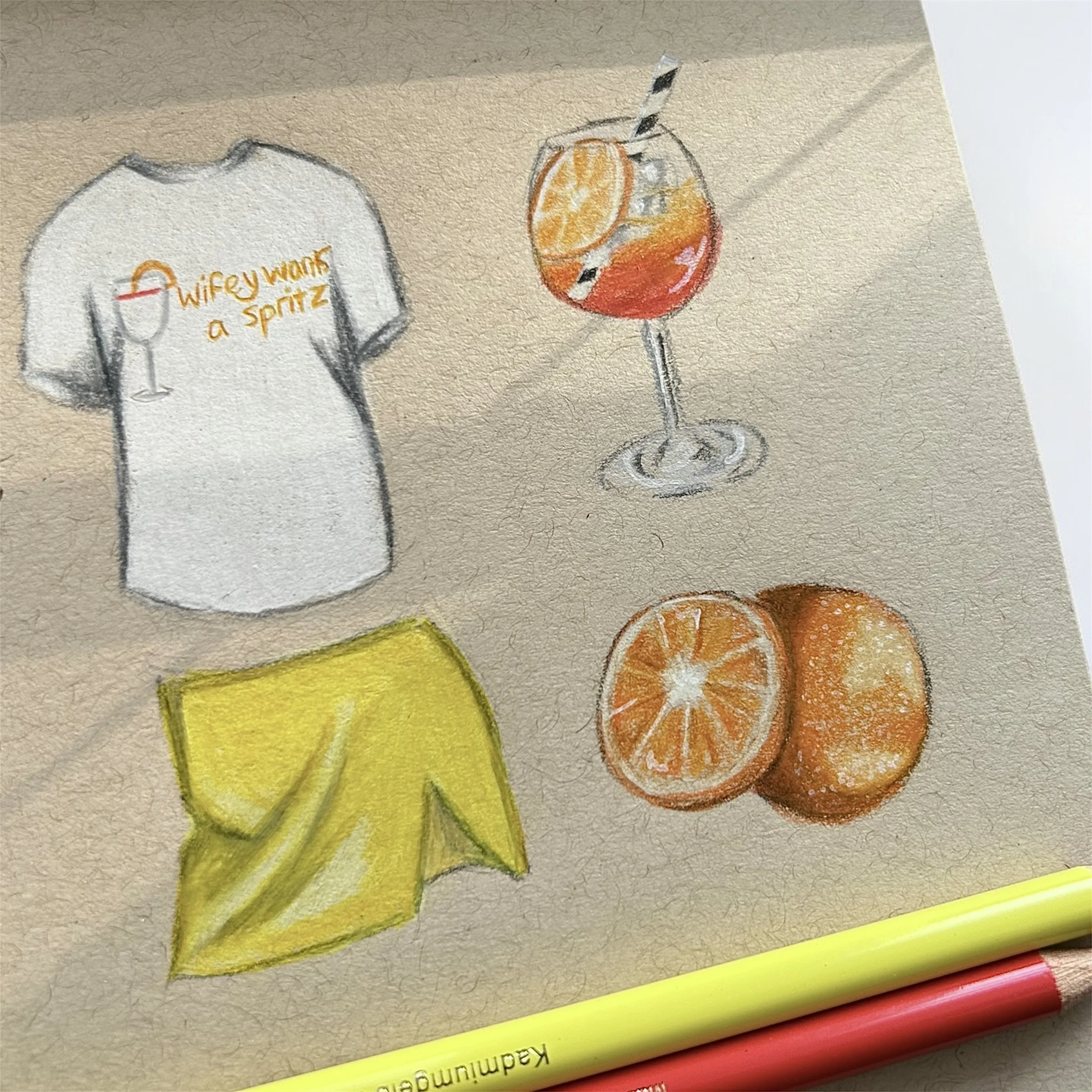 A hand-drawn illustration on tan paper featuring a white T-shirt with the text 'wifey wants a spritz' and a wine glass graphic, two oranges, a glass of red orange spritz with ice and a lemon slice, and a yellow cloth or napkin. There are also a yellow pencil and a red pencil at the bottom of the image.
