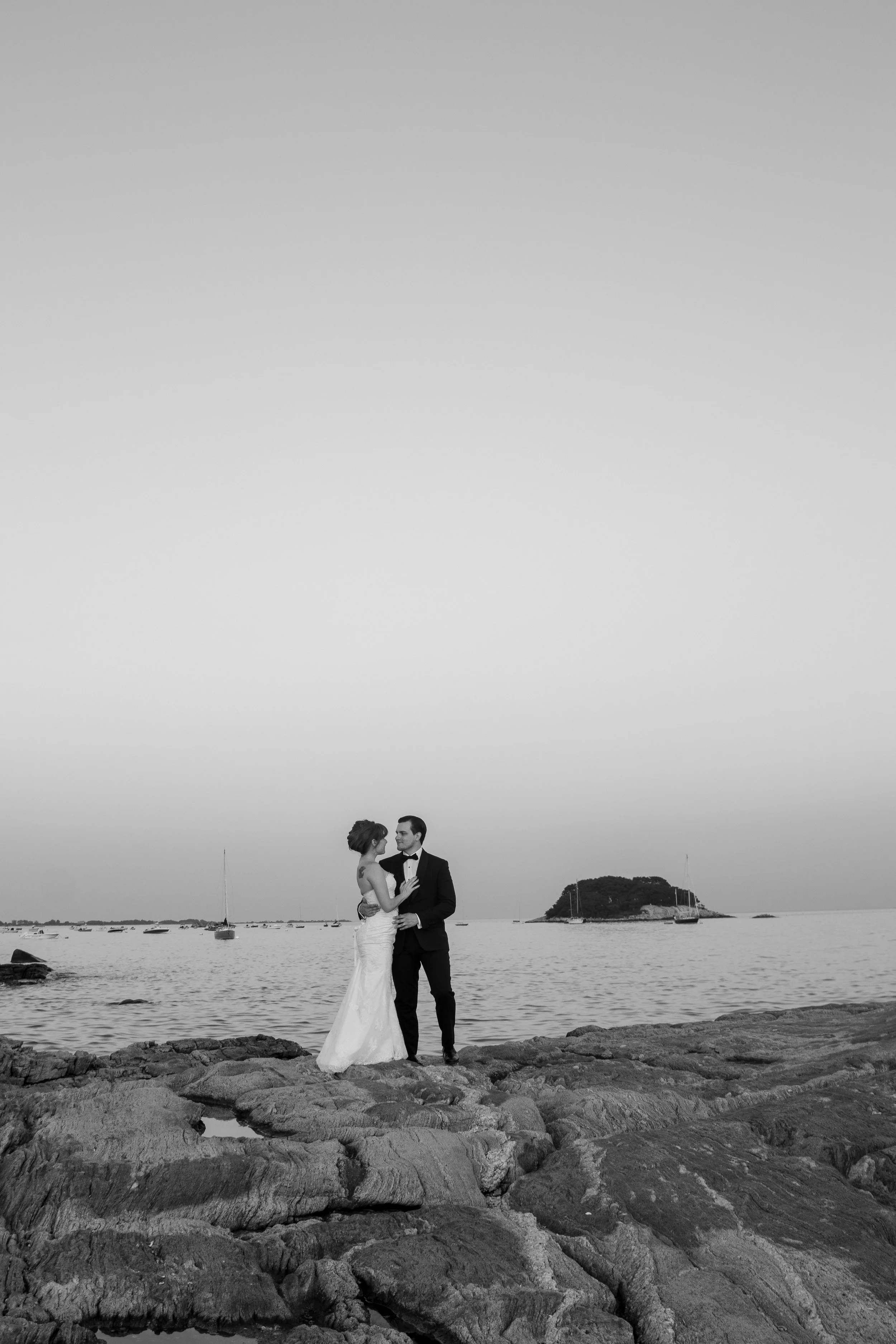 A black and white photograph of a bride and groom standing on rocks by the water, holding each other and looking at each other, with small boats and an island in the background.