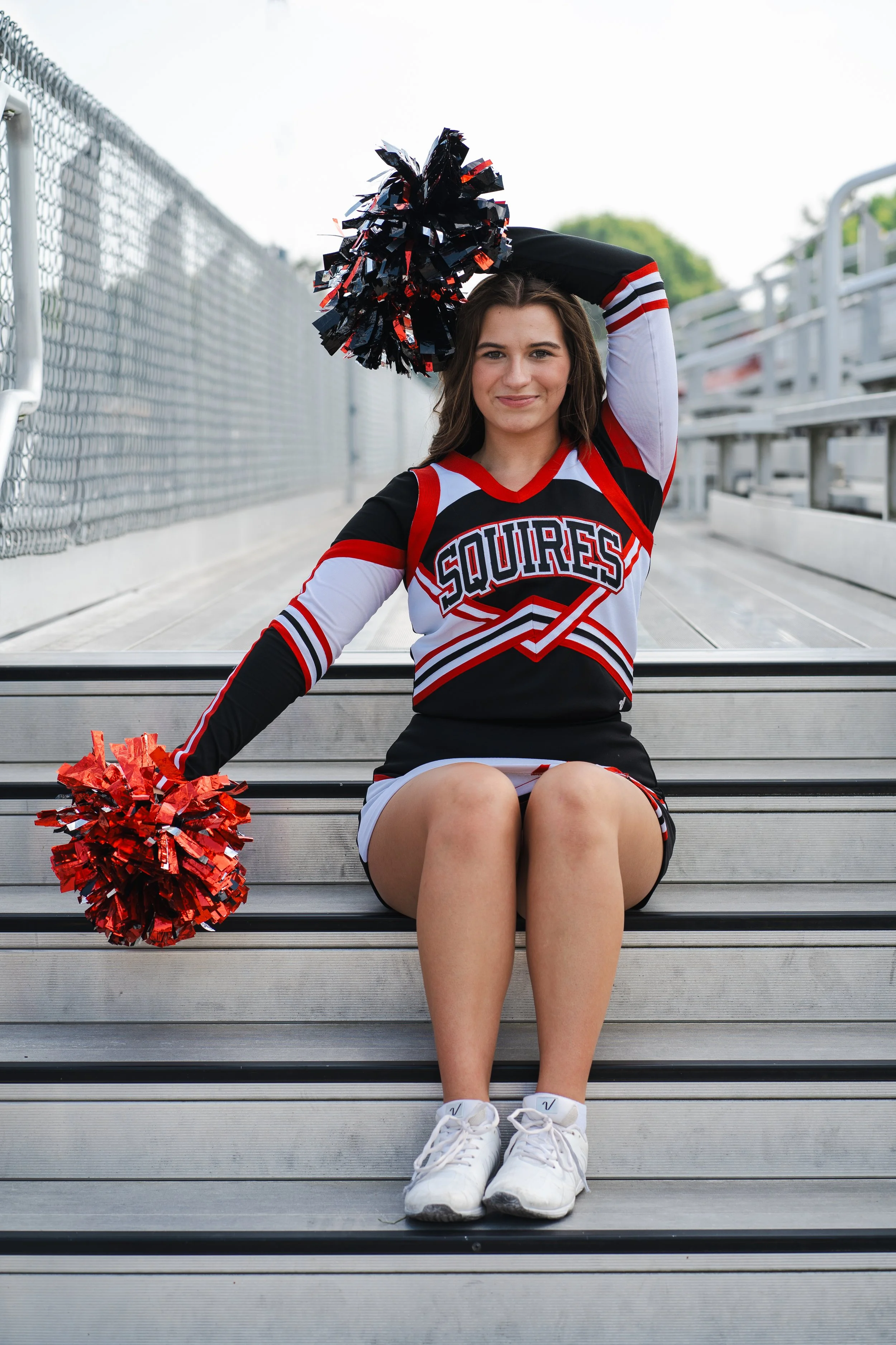 A cheerleader sitting on stadium bleachers, holding pom-poms, wearing a team uniform with 'Squires' on it.