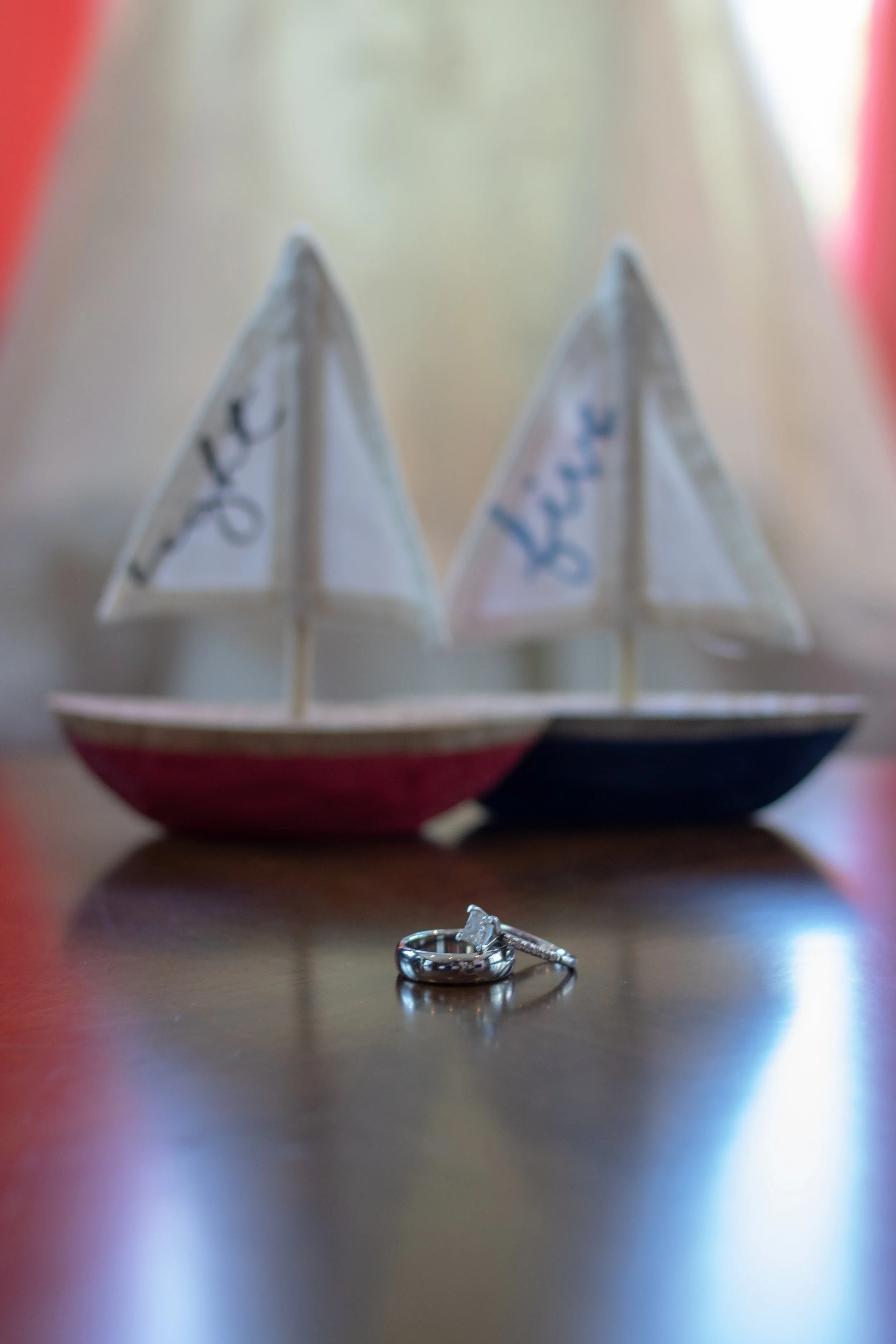 Wedding rings placed on a wooden surface with two small sailboat models in the background, one with the words "Husband" and "Wife" on the sails.