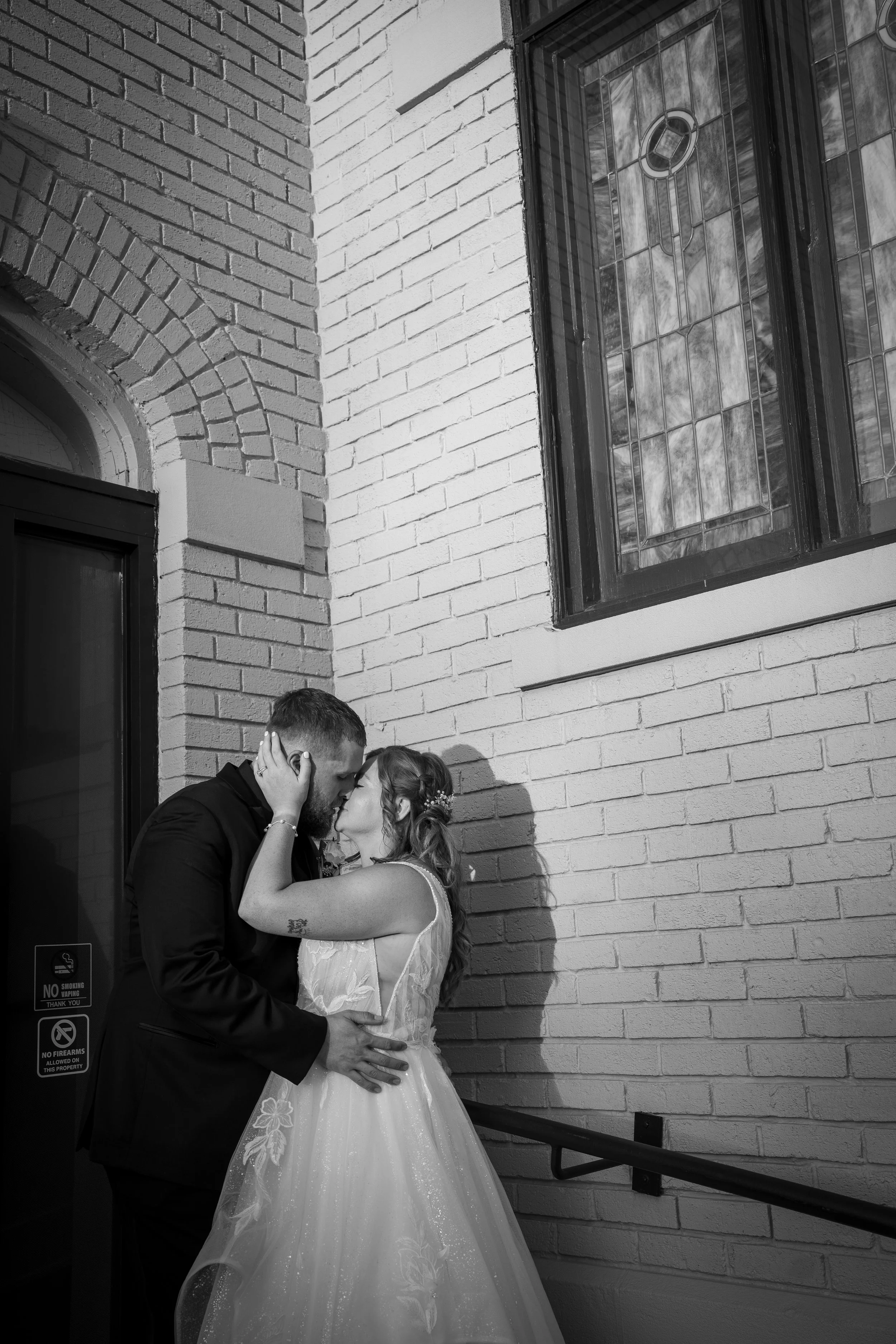A bride and groom in wedding attire sharing a kiss outside a building with brick walls and a stained glass window.