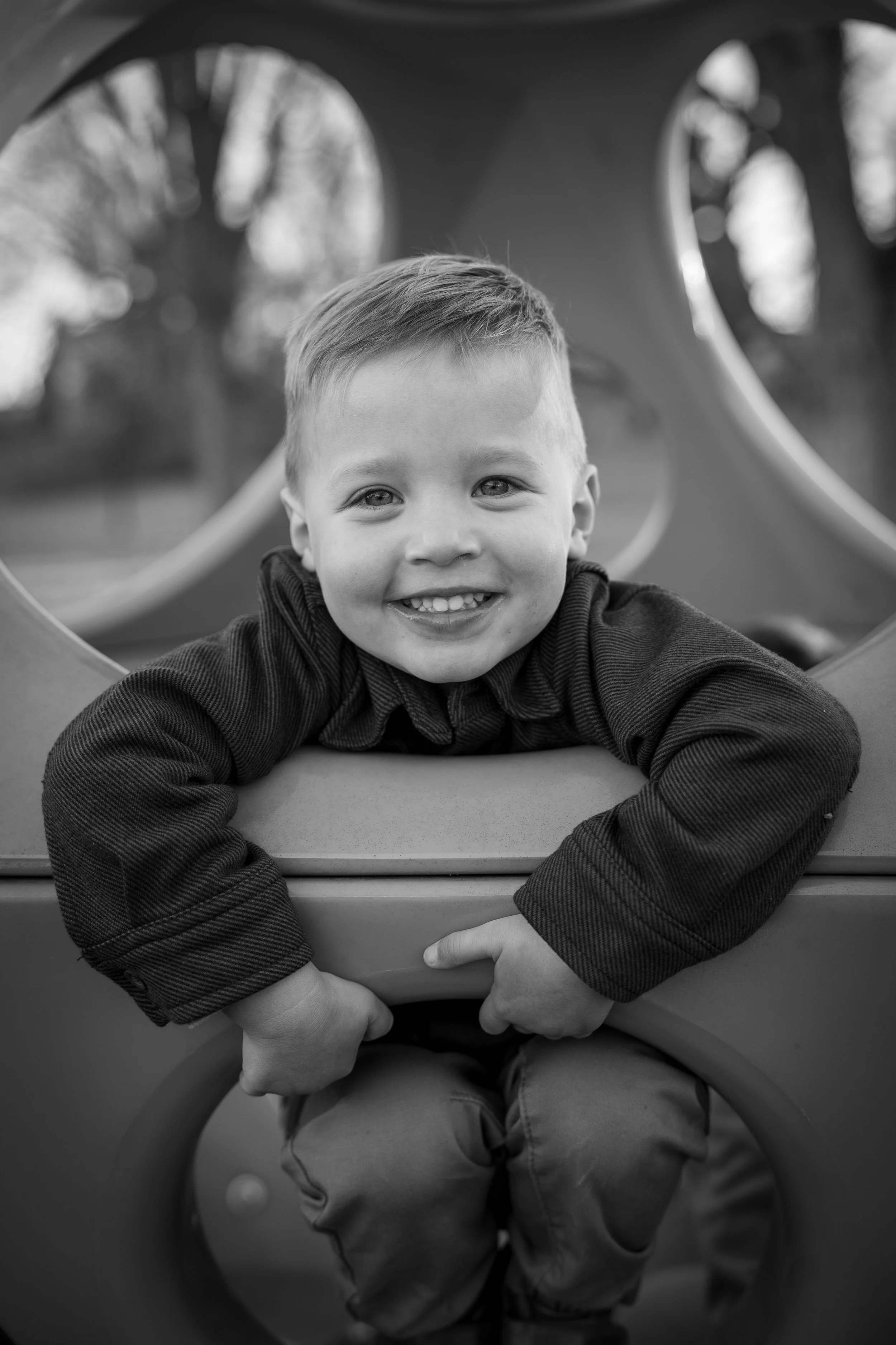 A smiling young boy leaning on a playground structure in an outdoor park, black and white photograph.