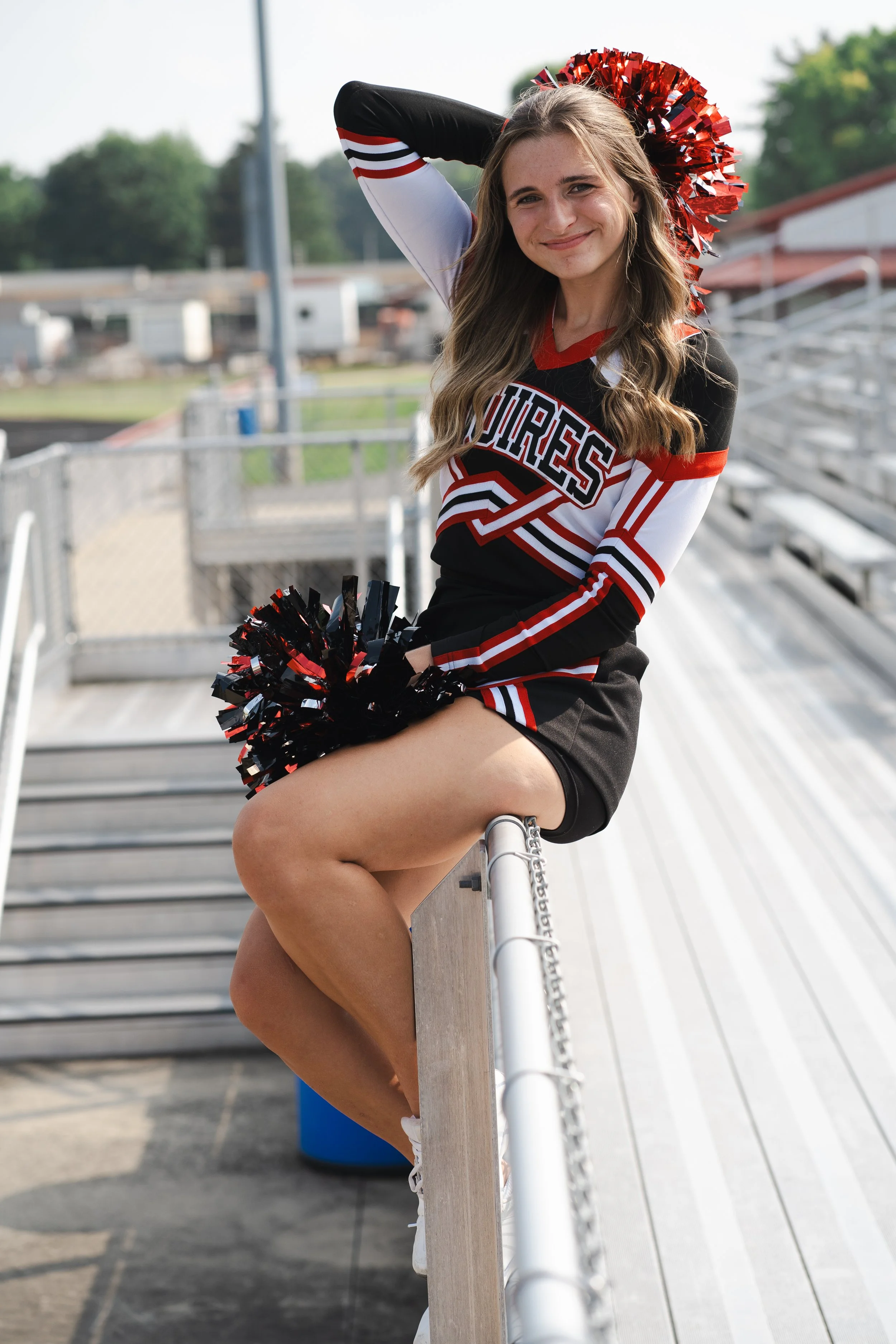 A young cheerleader sitting on a railing at a sports stadium, wearing a black, red, and white cheerleading uniform with pom-poms in her lap, and a red pom-pom hat, smiling at the camera.