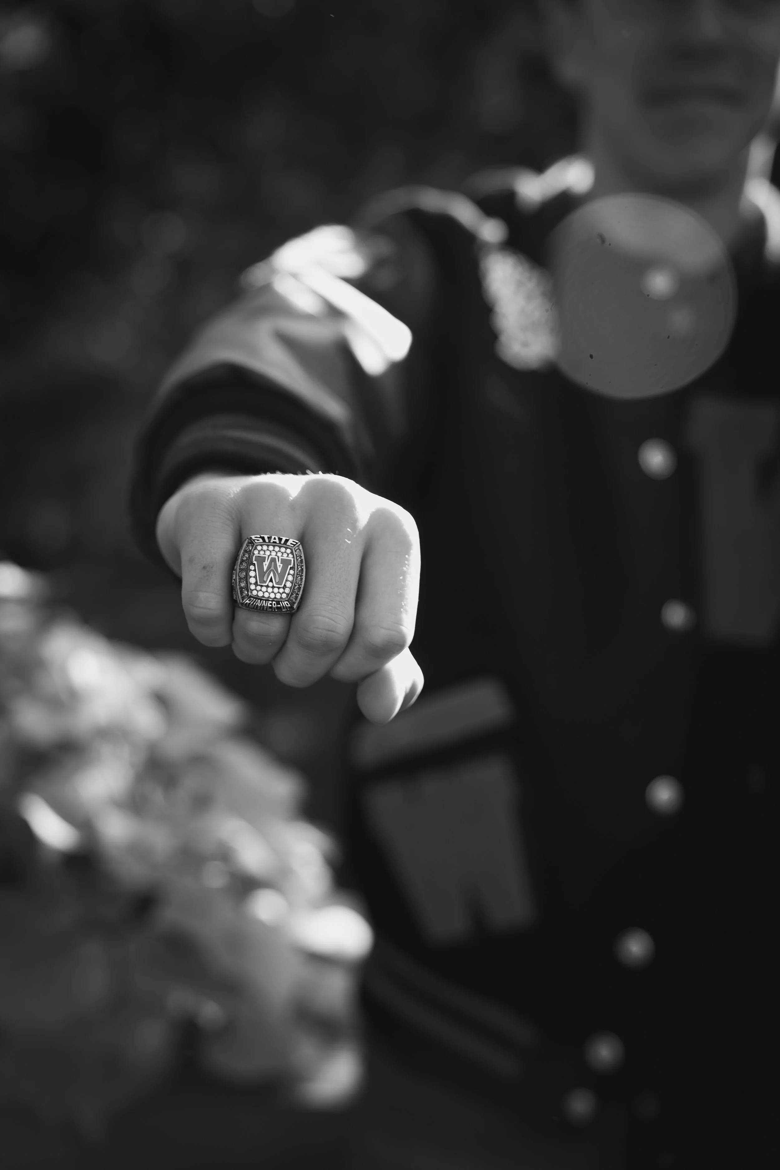 Close-up of a person's hand wearing a ring with a sports team logo, with the person making a fist toward the camera, in black and white.