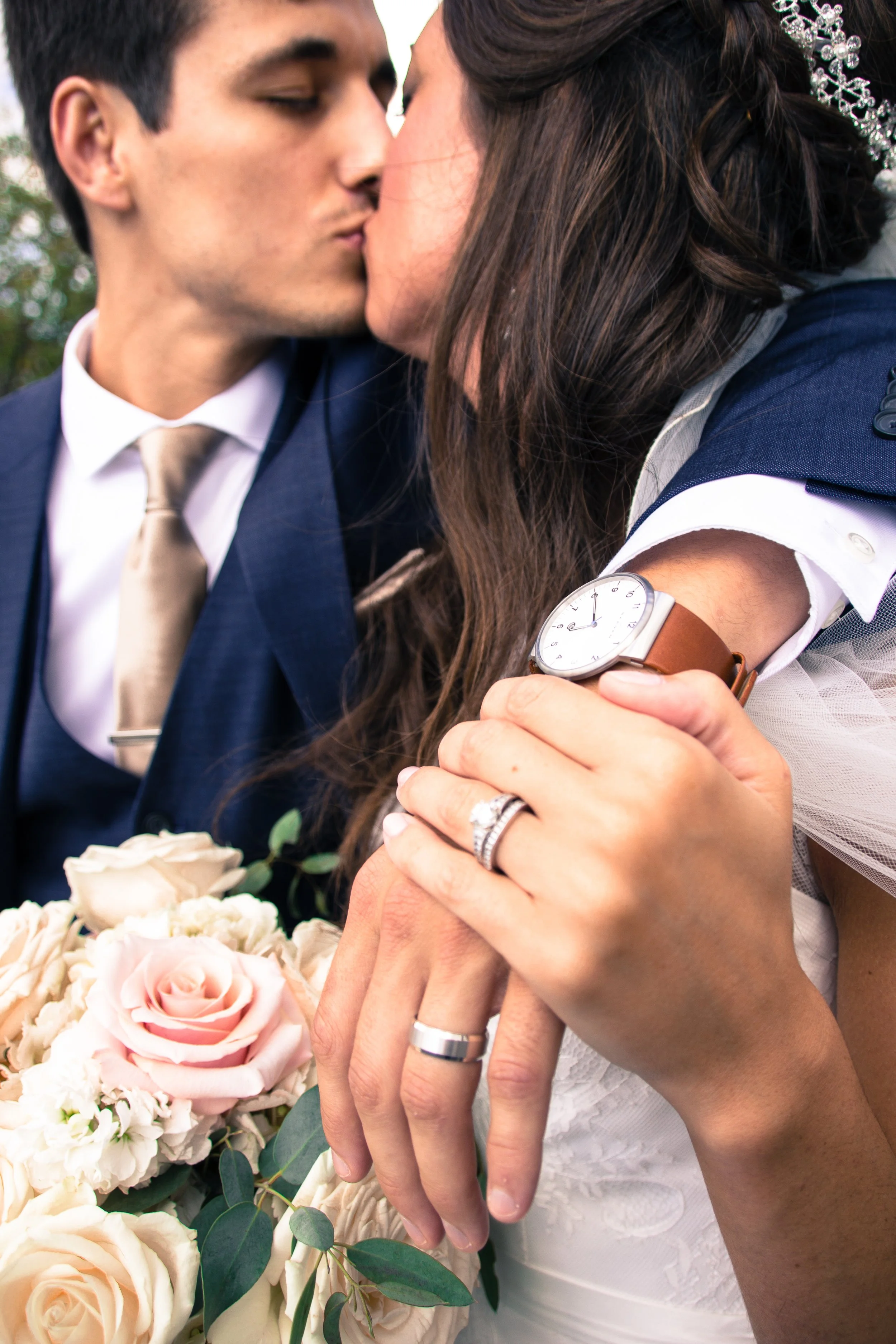 A newly married couple sharing a kiss, with the bride holding a bouquet of pink and white roses, and their hands showing wedding rings, outdoors with greenery in the background.