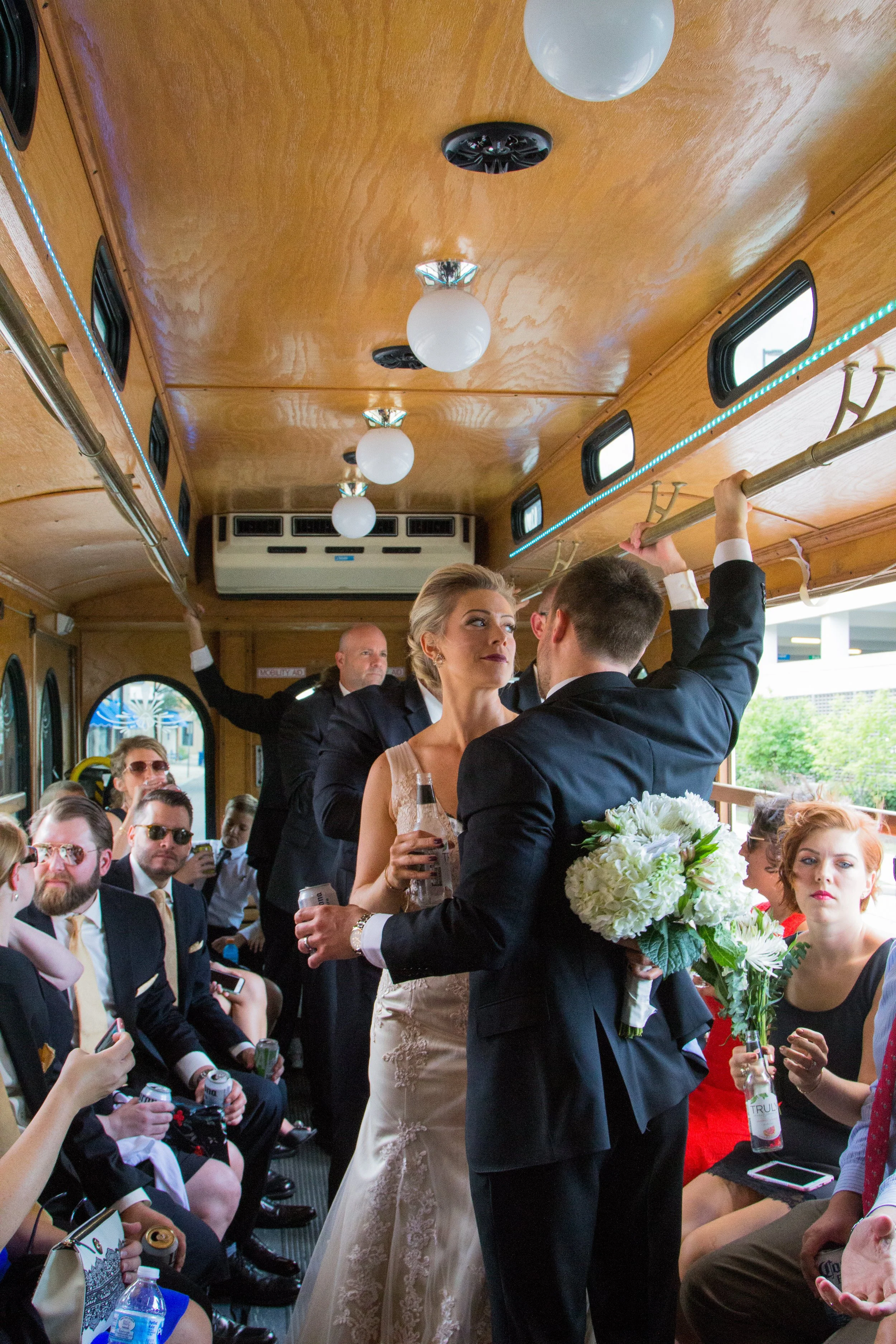 A wedding celebration inside a bus or trolley with couples dancing and guests sitting, some holding drinks and wearing formal attire.