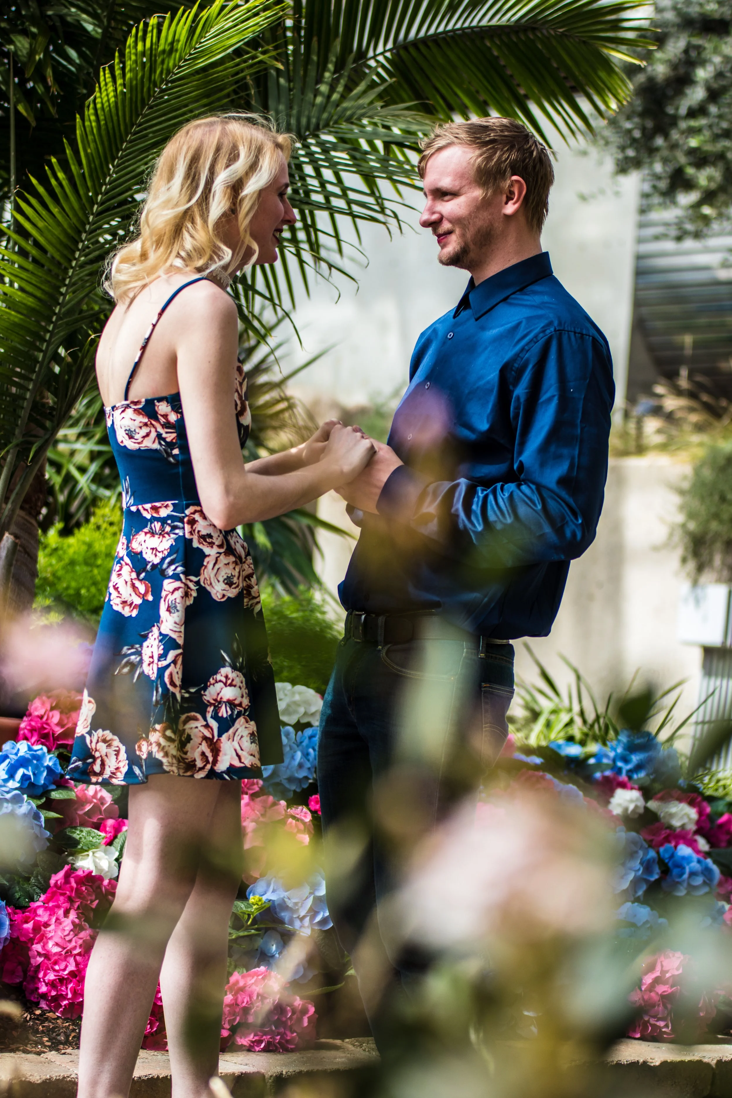 A couple holding hands and gazing at each other in a garden with colorful flowers and lush green plants.