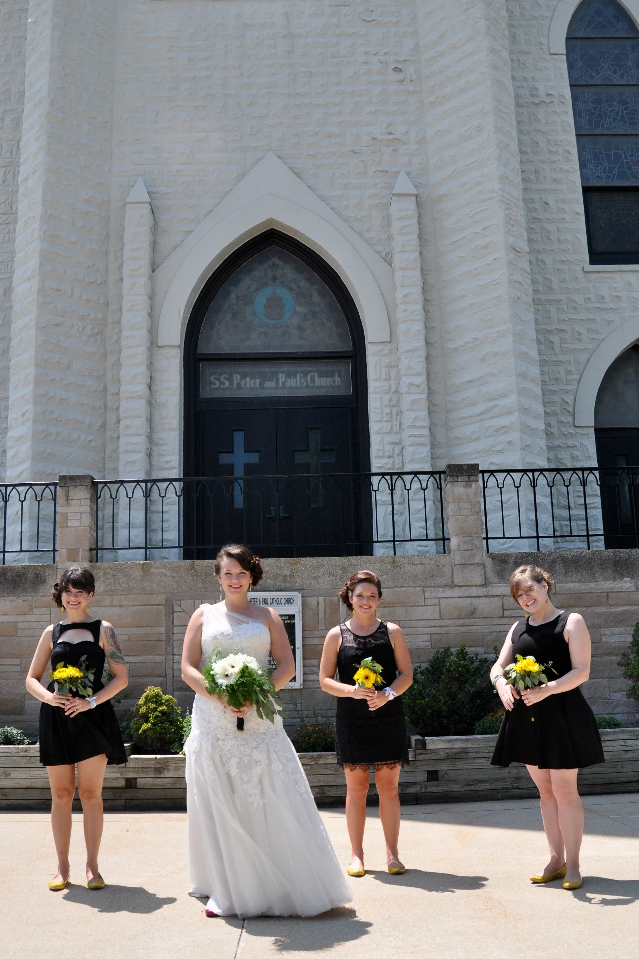 A bride and three bridesmaids standing outside a white church. The bride is in a white wedding gown holding a bouquet, and the bridesmaids in black dresses holding yellow and white flowers.