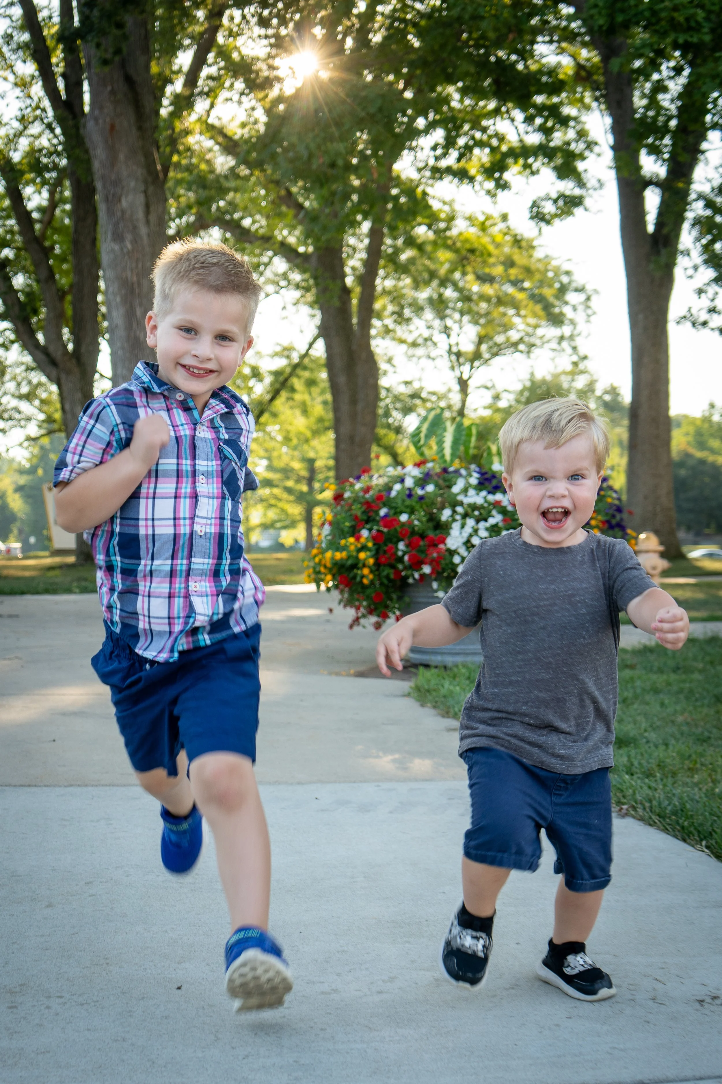Two young boys running on a sidewalk in a park, with trees and a flower pot in the background, sunlight shining through the trees.