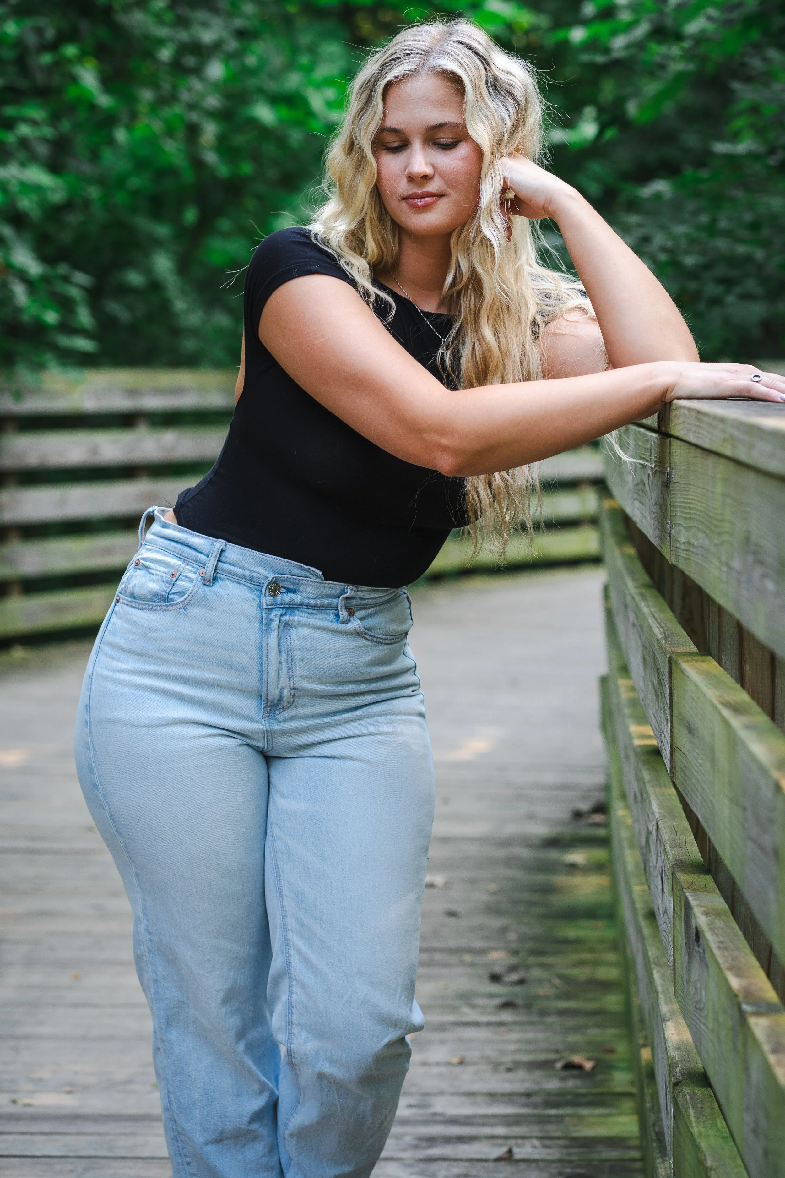 A woman with long wavy blonde hair, wearing a black top and light blue jeans, leaning on a wooden railing outdoors surrounded by greenery.