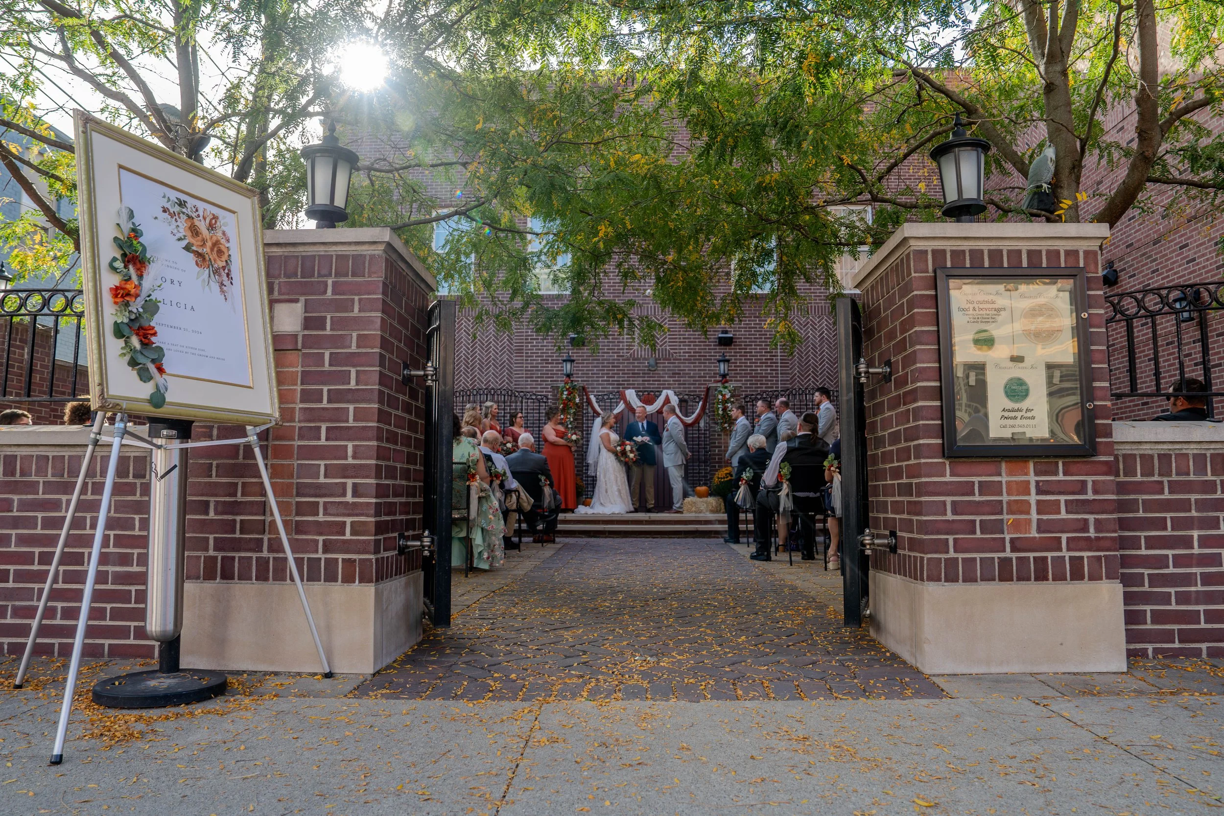 Outdoor wedding ceremony with bride and groom exchanging vows in front of officiant, seated guests, decorative floral arrangements, and banners, under a tree with sunlight filtering through.