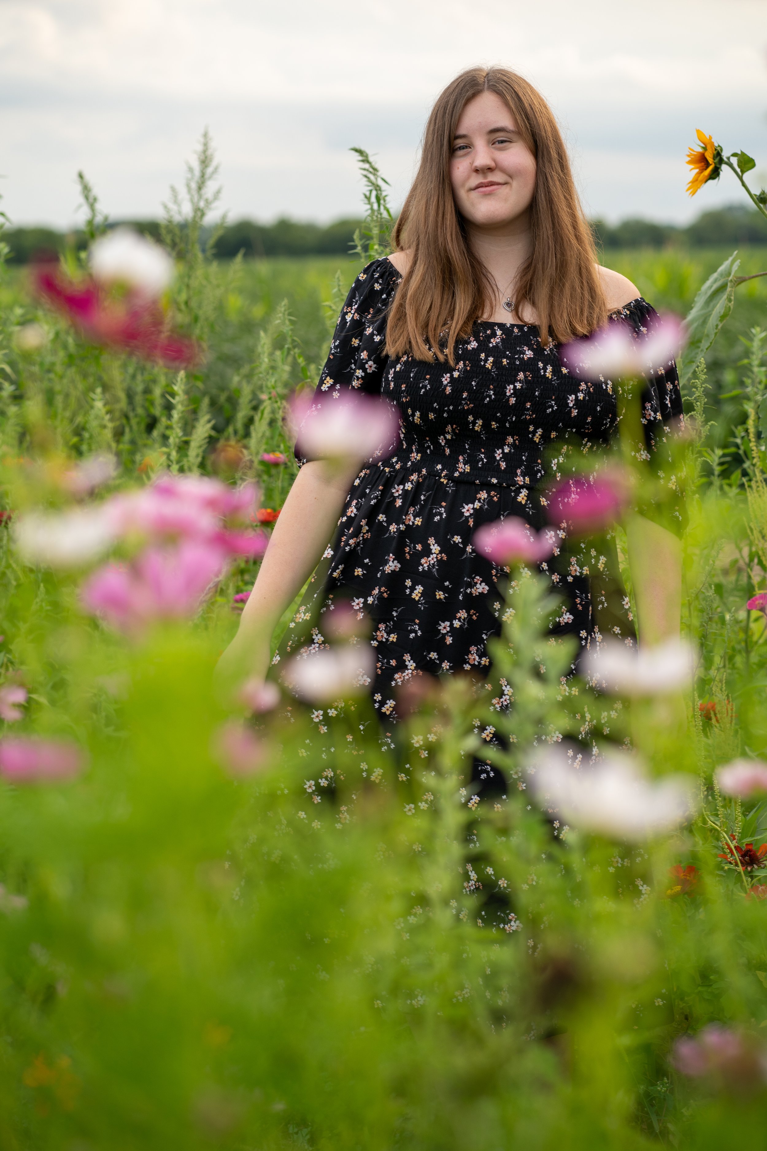 Young woman with brown hair wearing a black floral dress standing in a field of wildflowers under a cloudy sky.