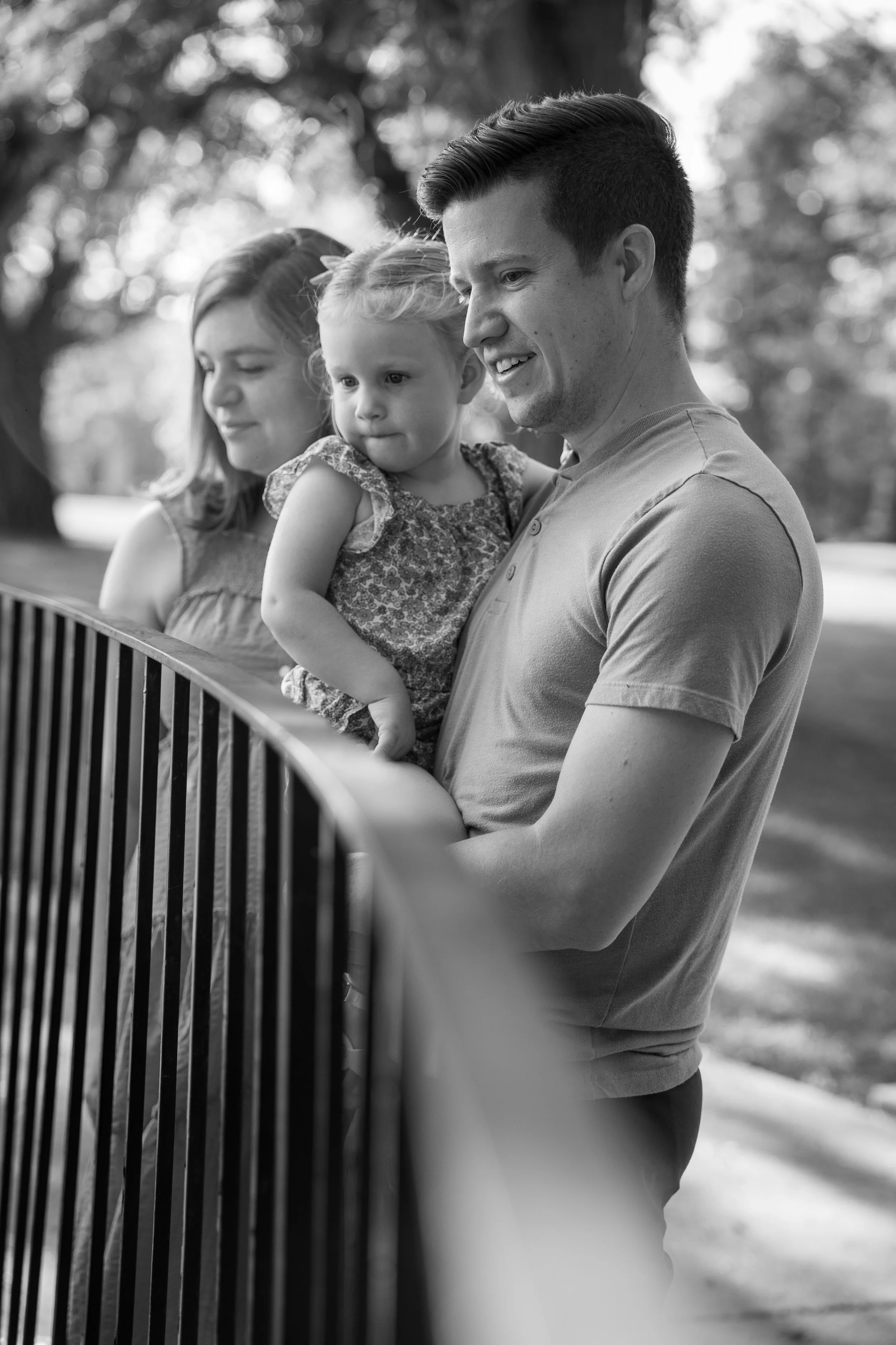 A man holding a young girl, with a woman beside them, all looking at something beyond the camera in a park setting, black and white photo.