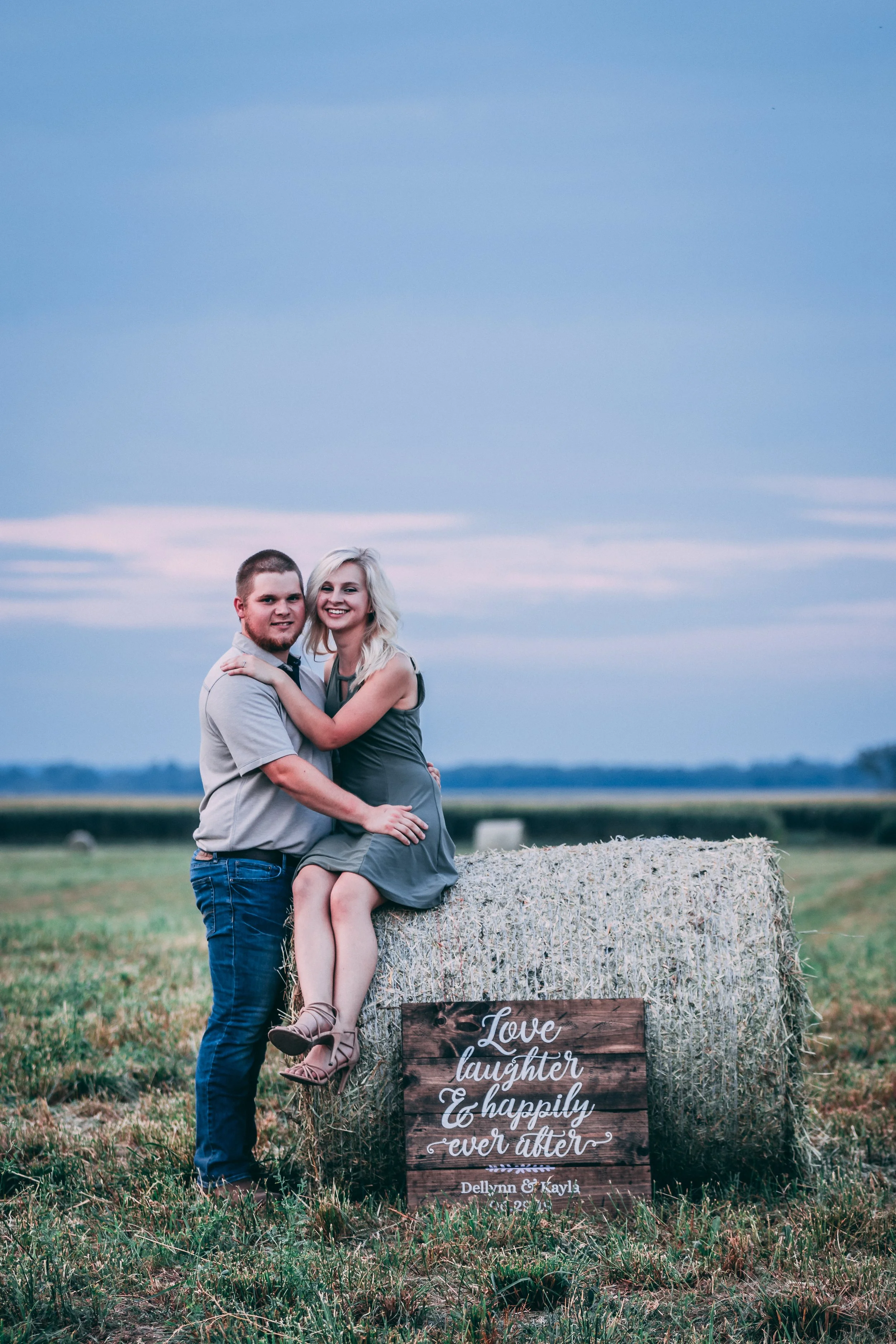 A smiling couple with the woman sitting on a hay bale and the man standing next to her, outdoors in a field with a sign that reads 'Love, laughter & happily ever after, Delynn & Kayla, 09-12-20'.