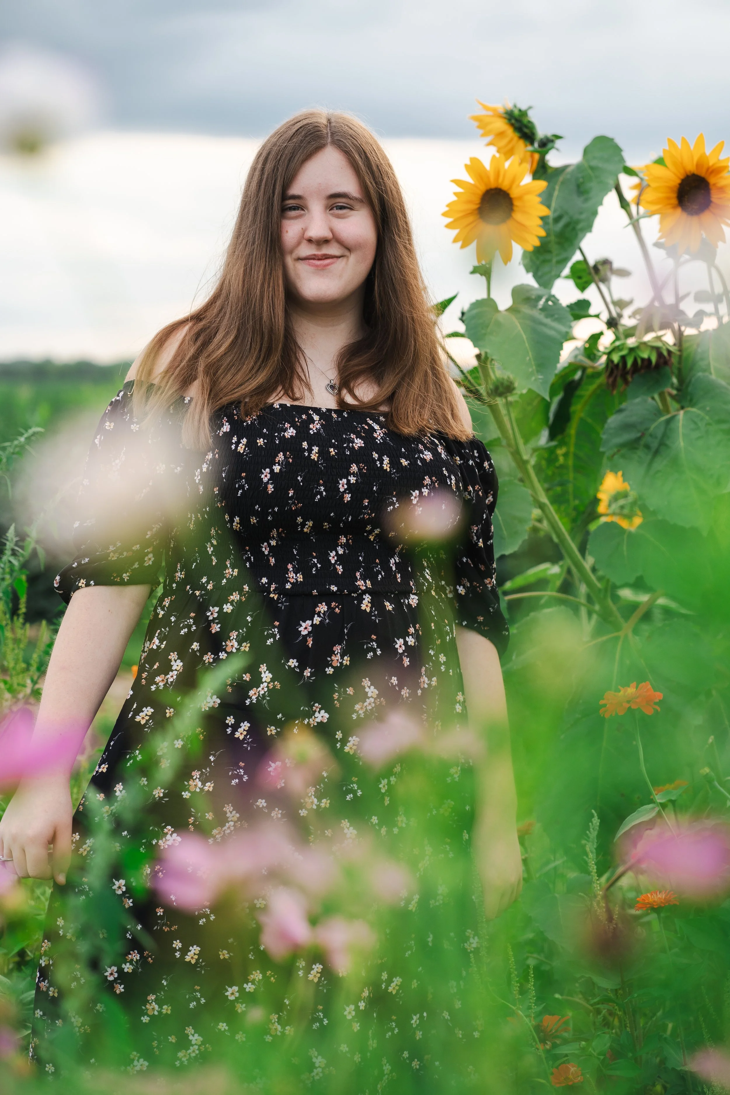A young woman with long brown hair wearing a black floral dress standing among sunflowers and other colorful flowers in a garden or field.