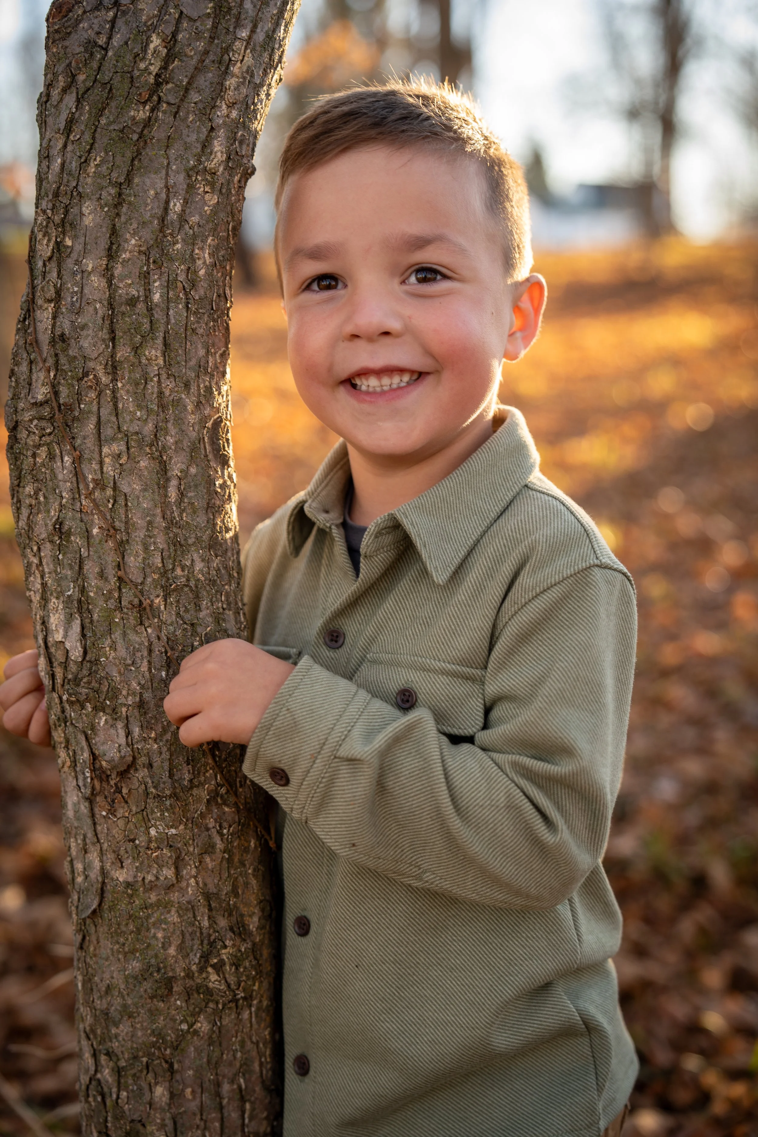 A young boy in a green shirt smiling and holding a tree in an outdoor park during autumn.