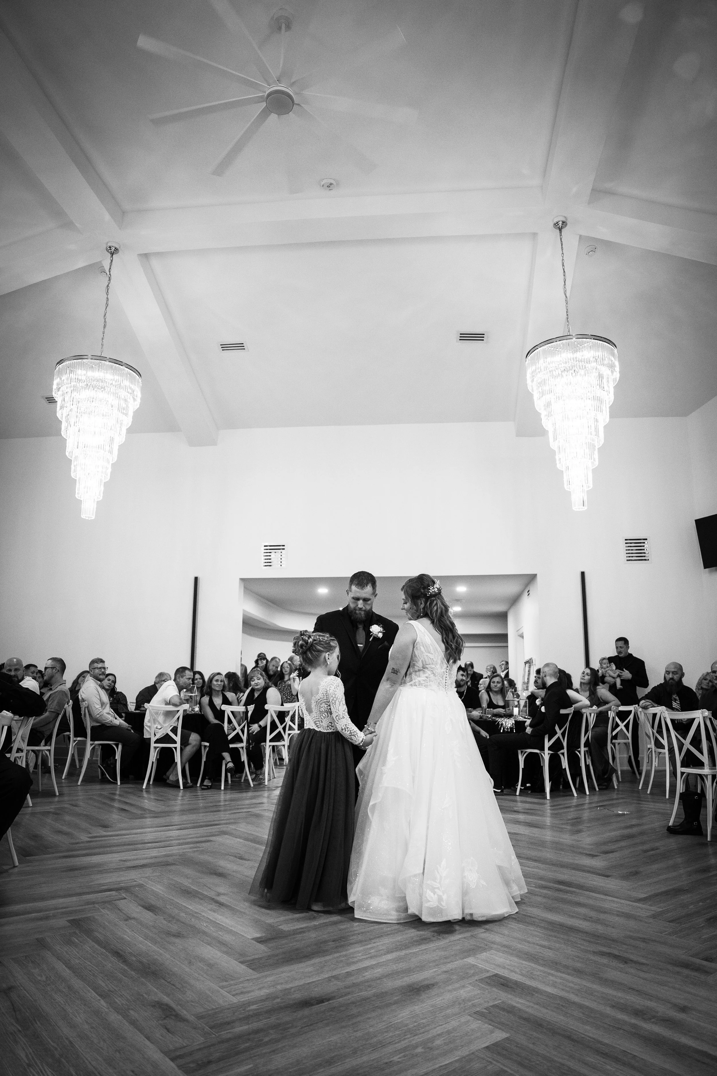 A bride and groom dance with a young girl at their wedding reception in a banquet hall, with guests seated at tables watching.