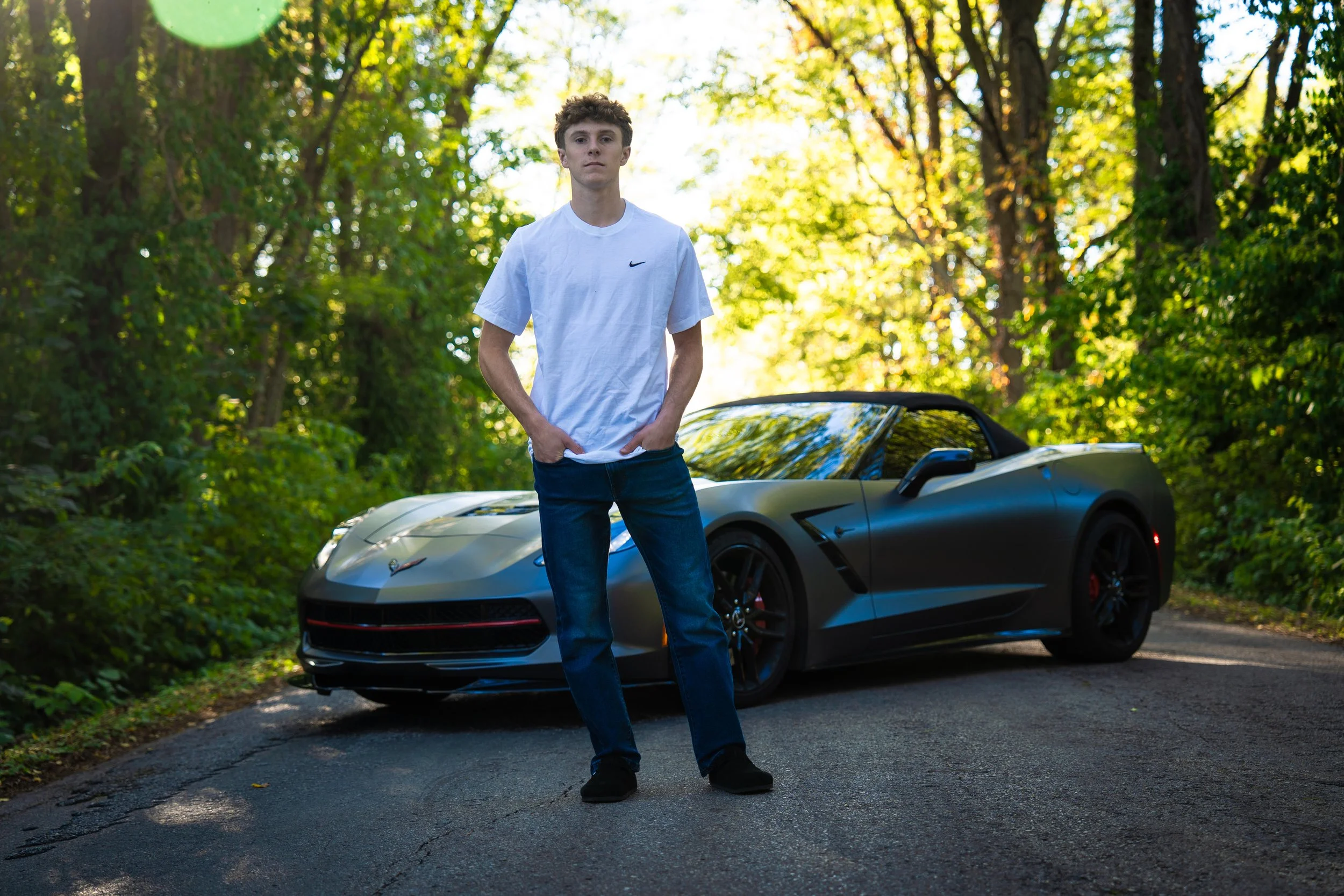 Young man in white t-shirt and jeans standing in front of a black sports car on a forested road.