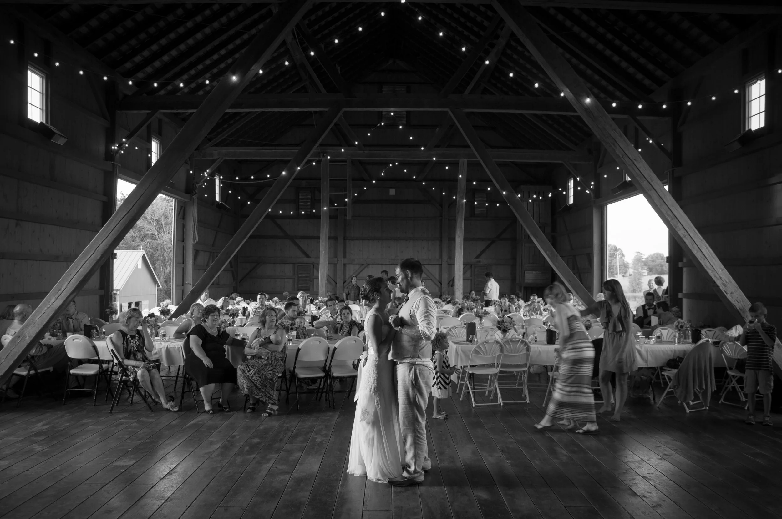 A black and white photograph of a wedding reception inside a rustic barn. The bride and groom are dancing together in the center. Guests are seated and standing around tables decorated with flowers. String lights are hanging from the ceiling.
