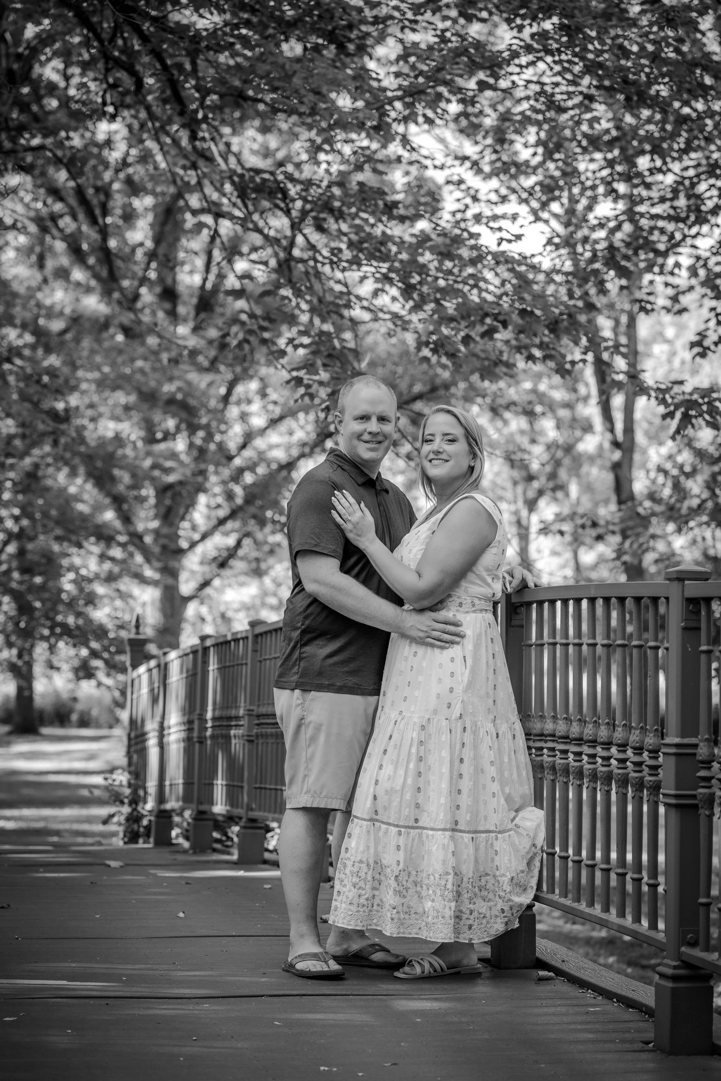 Black and white photo of a couple standing on a wooden bridge surrounded by trees, smiling and embracing each other.