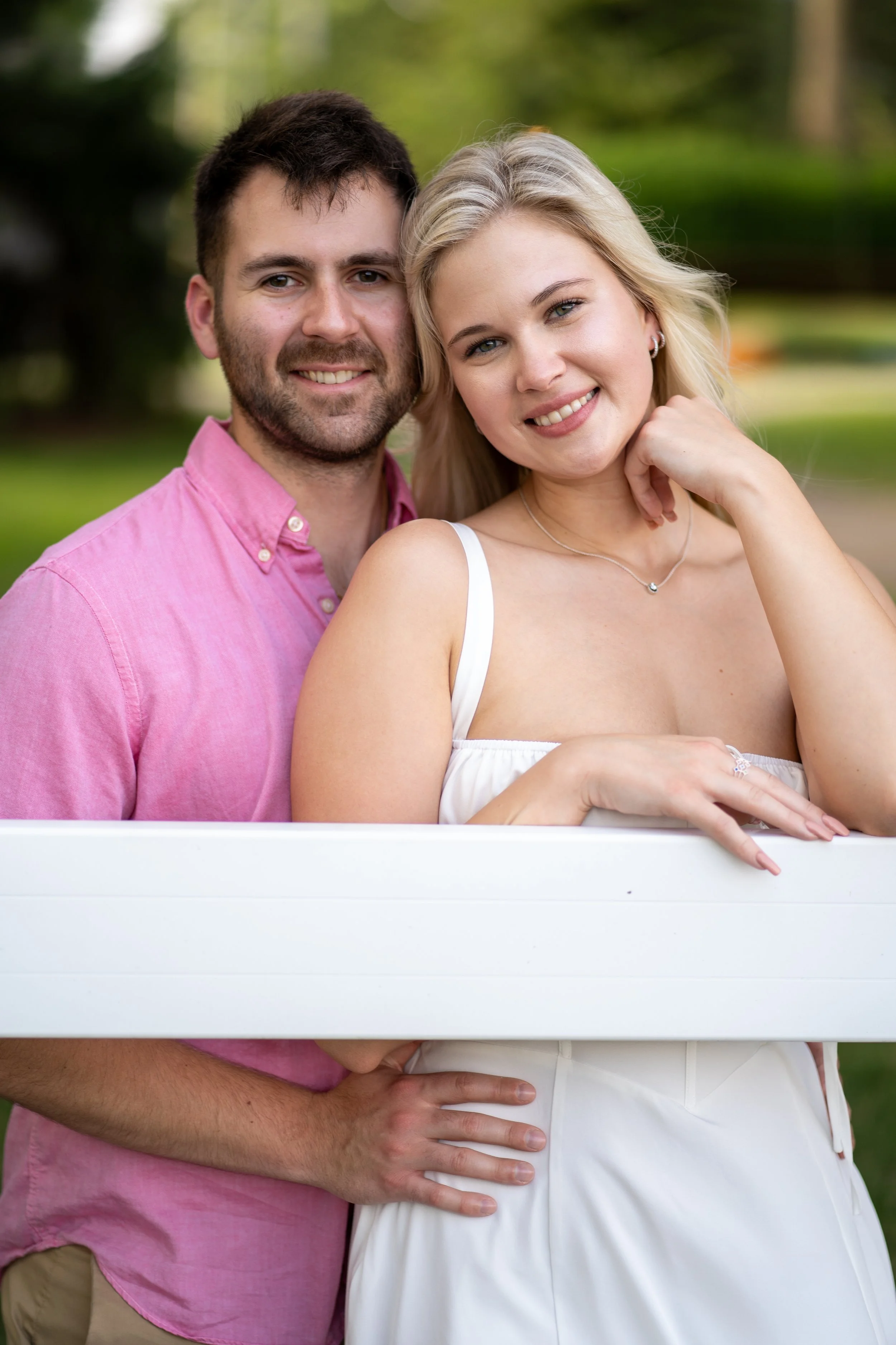 A smiling couple, a man in a pink shirt and a woman in a white dress, standing outdoors in a park with trees and grass in the background.
