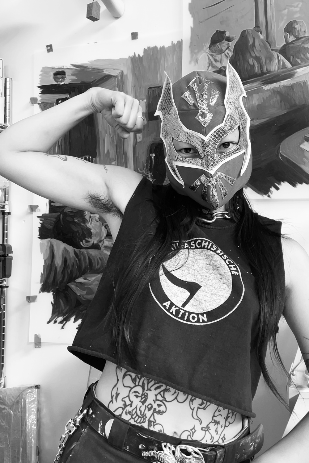 A person wearing a lucha libre mask, a sleeveless t-shirt with the logos and text, and a belt, flexing their right arm in front of a painted background.