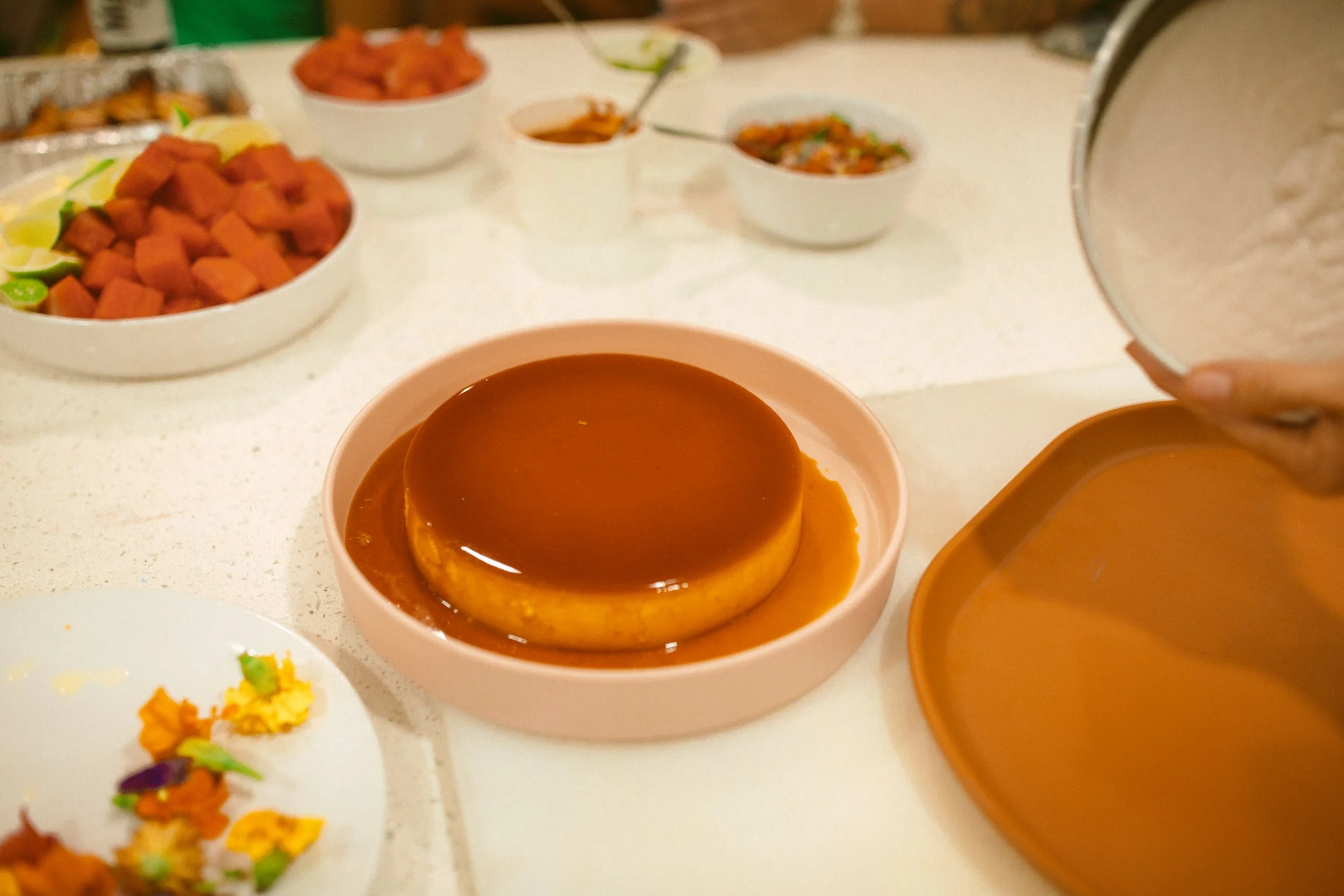Close-up of a caramel flan dessert on a pink plate with caramel sauce on top, surrounded by various dishes including bowls of watermelon, fruit, and other foods on a white table.
