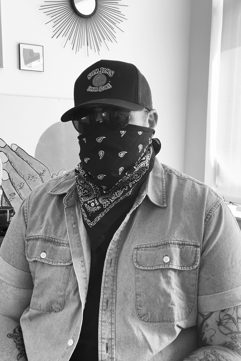 Person wearing a cap, sunglasses, and a bandana covering their face, sitting indoors with a wall and window behind them.