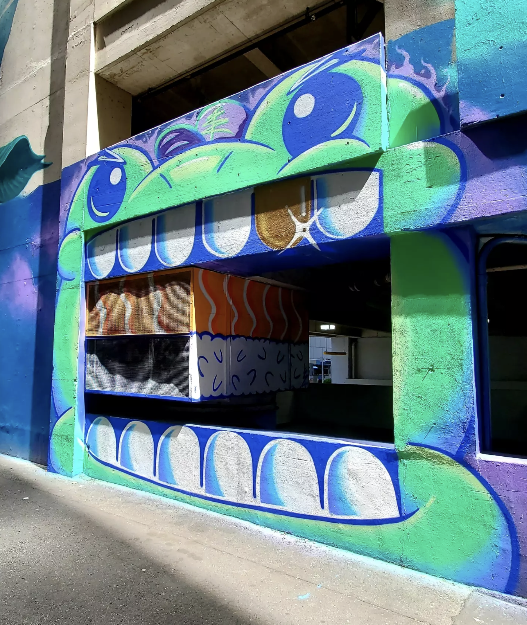 Colorful street art mural of a large green and blue creature with big eyes and a wide smile painted on a building facade.