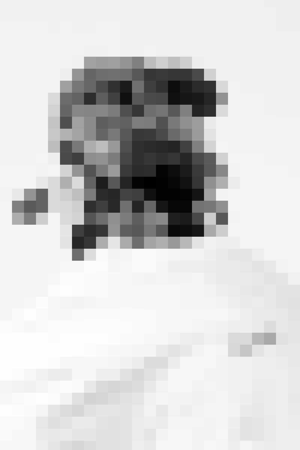A pixelated black and white photo of a person with a beard, wearing glasses and a hat, sitting at a table.
