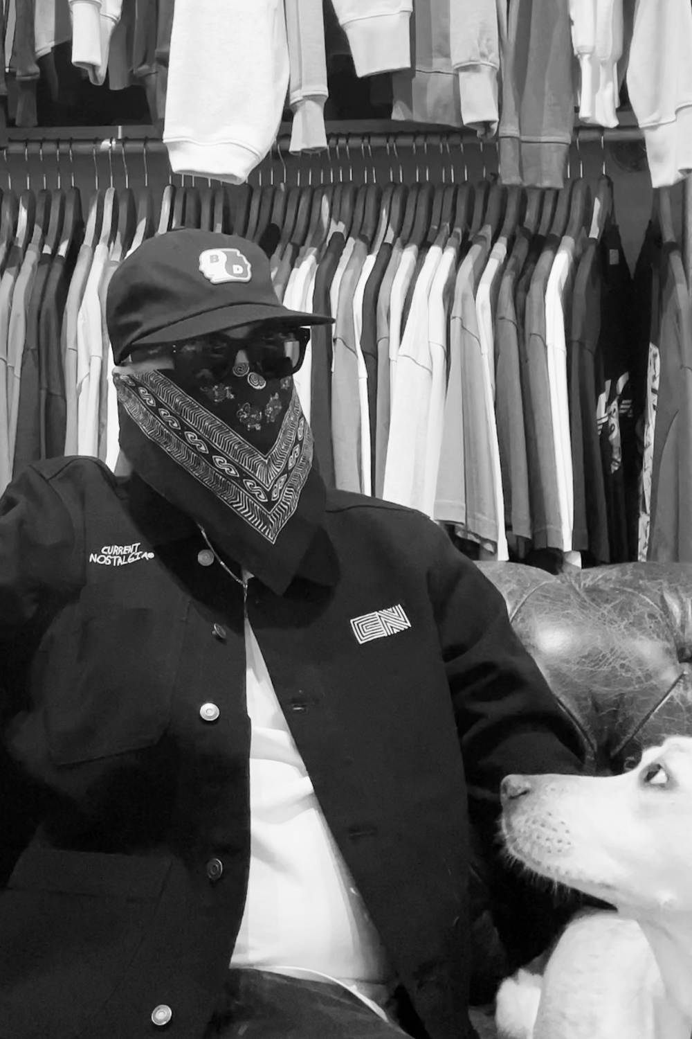 A person wearing a baseball cap, sunglasses, bandana, and black jacket sitting on a leather couch with a dog nearby, in front of a clothing rack filled with hanging shirts.