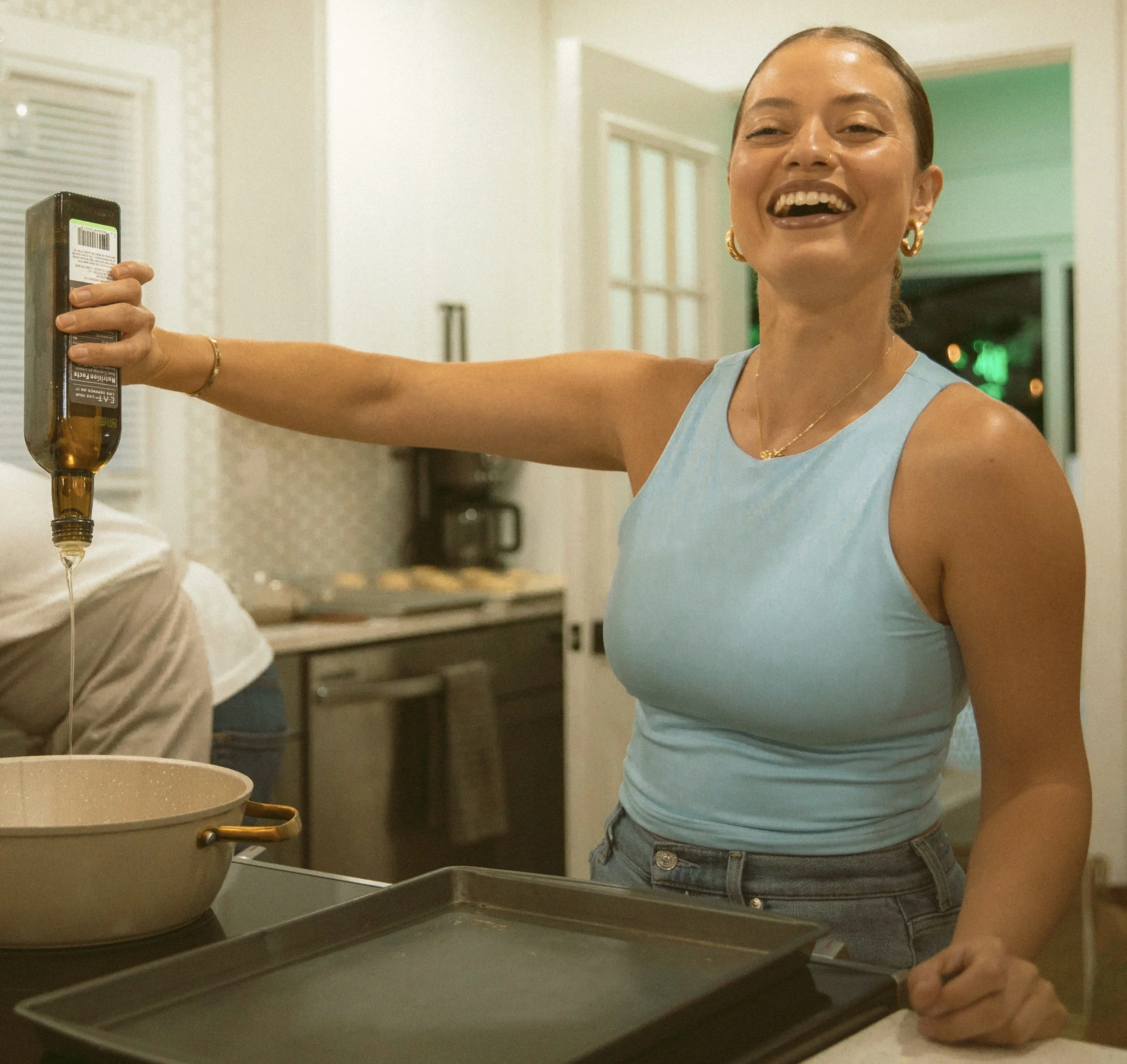 A woman in a light blue sleeveless top and jeans pouring oil into a baking dish in a kitchen, smiling.